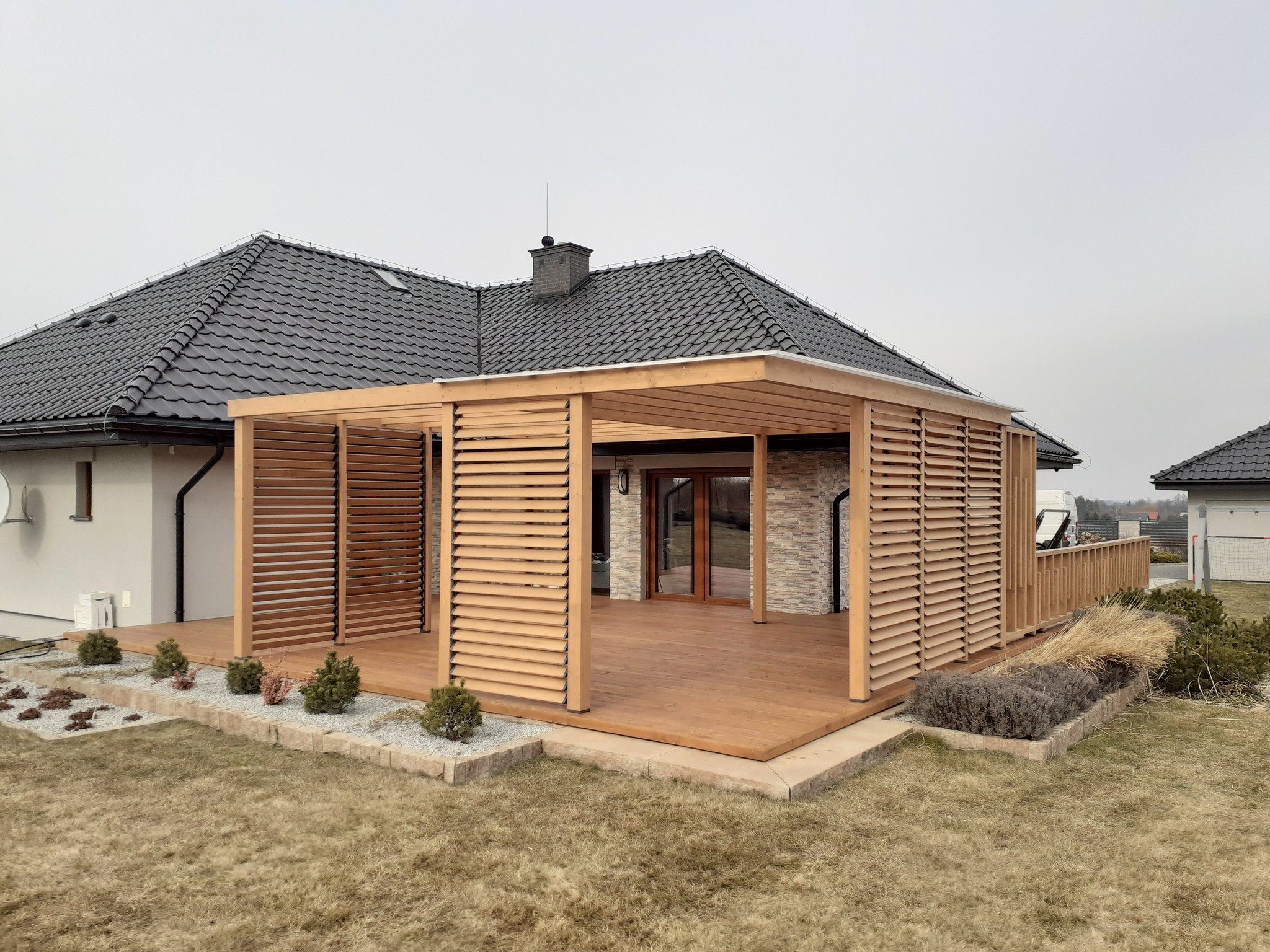 Drewniana konstrukcja z żaluzjami osłaniająca taras przy nowoczesnym domu z szarą dachówką, otoczona trawnikiem i ozdobnymi krzewami.