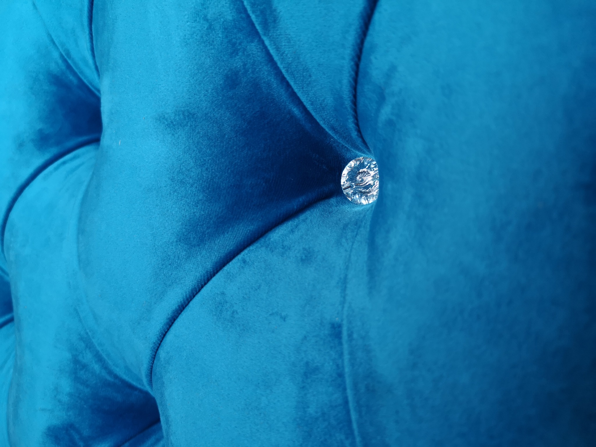 Fragment pikowanego zagłówka łóżka obitego turkusowym welurem, ozdobionego kryształowym guzikiem.