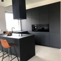 Nowoczesna, minimalistyczna kuchnia w kolorze grafitowym z wyspą i skórzanymi hokerami, oświetlona naturalnym światłem z okna.