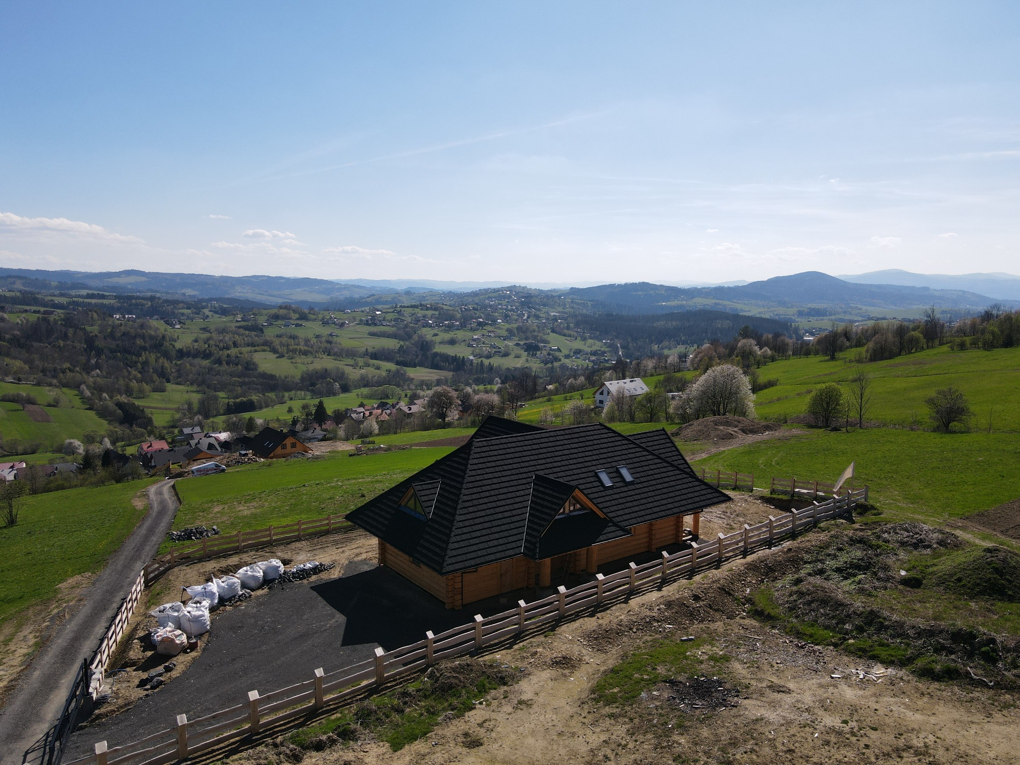 Drewniany dom z bali z czarnym dachem, widok z lotu ptaka na pagórkowaty krajobraz z elementami wsi i lasu, w tle błękitne niebo.