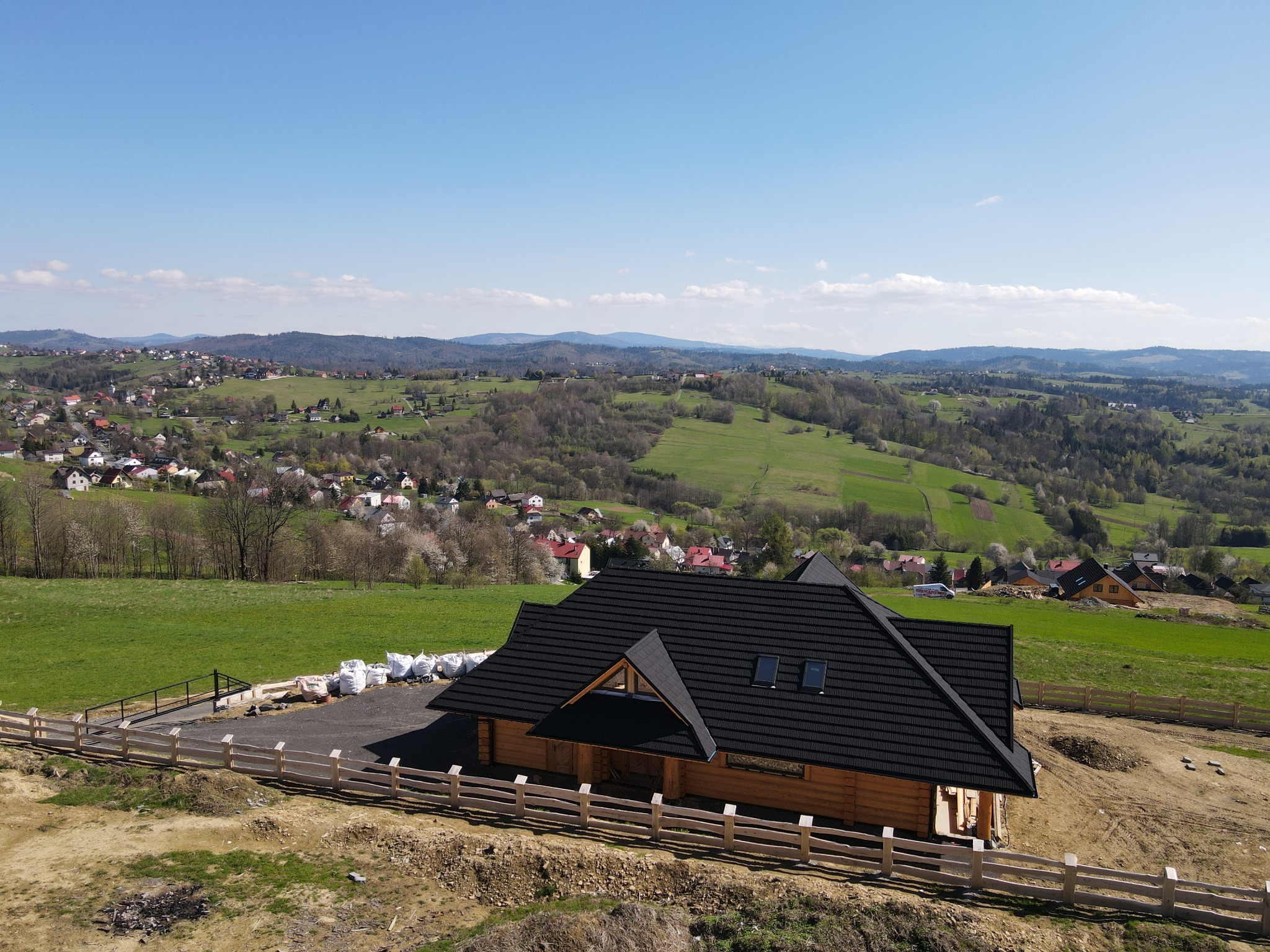 Nowo wybudowany, parterowy dom z bali z czarnym dachem na tle malowniczego krajobrazu wzgórz i pól, ogrodzony drewnianym płotem, z widocznymi workami materiałów budowlanych obok.