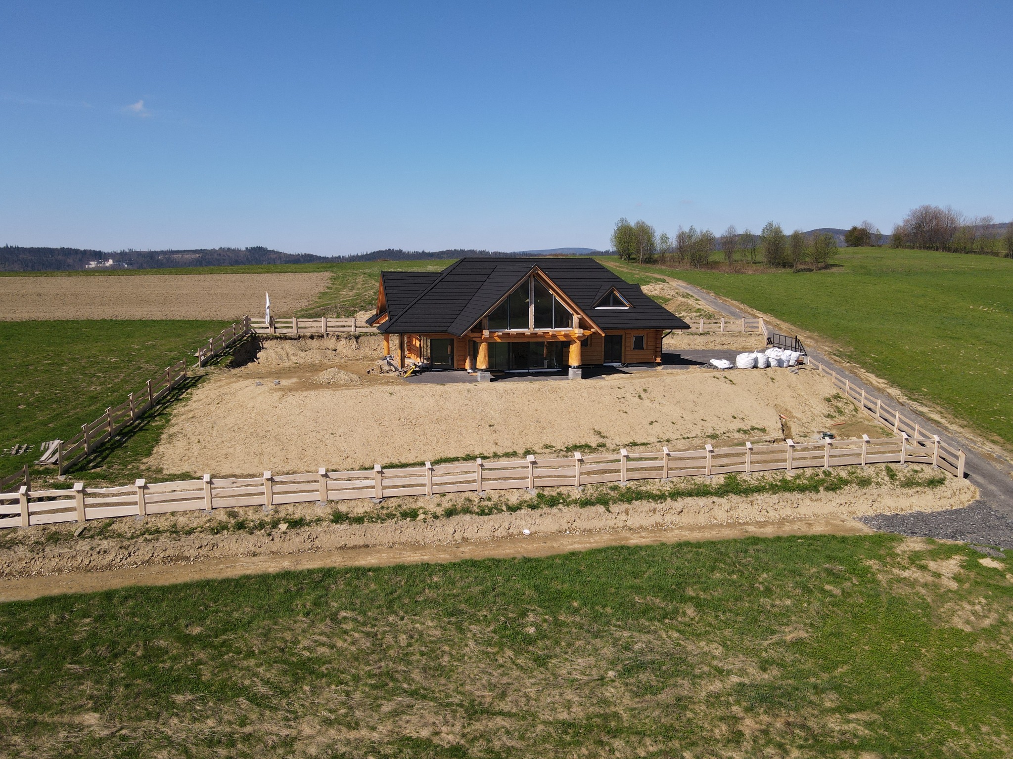 Nowoczesny dom z drewnianych bali z czarnym dachem, panoramicznymi oknami i drewnianym ogrodzeniem w trakcie budowy, widok z drona na pagórkowaty teren.
