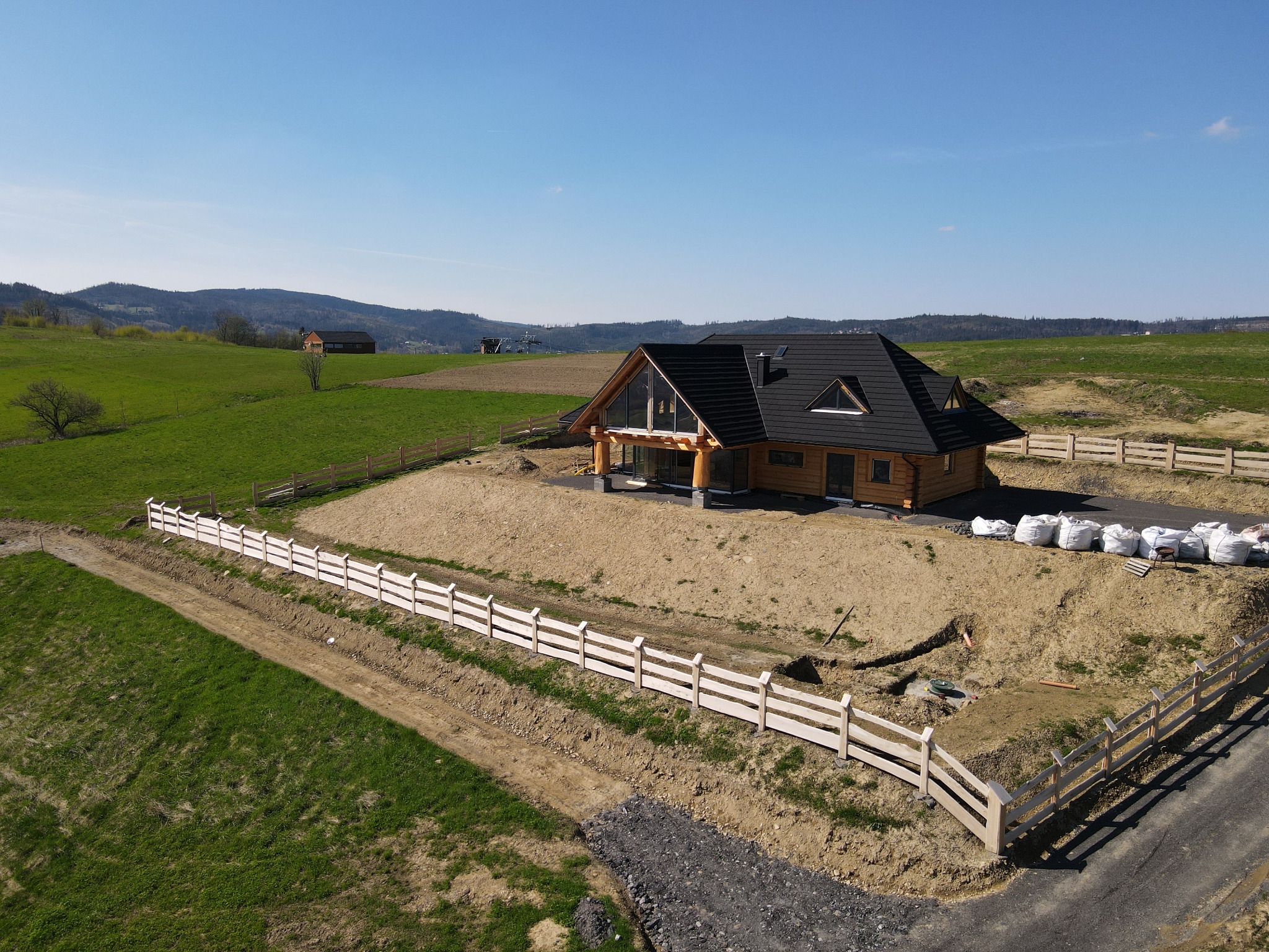 Nowoczesny, parterowy dom z bali z ciemnym dachem, widziany z lotu ptaka na tle zielonych wzgórz, ogrodzony jasnym, drewnianym płotem. Wokół domu widoczne worki z materiałami budowlanymi i świeżo...
