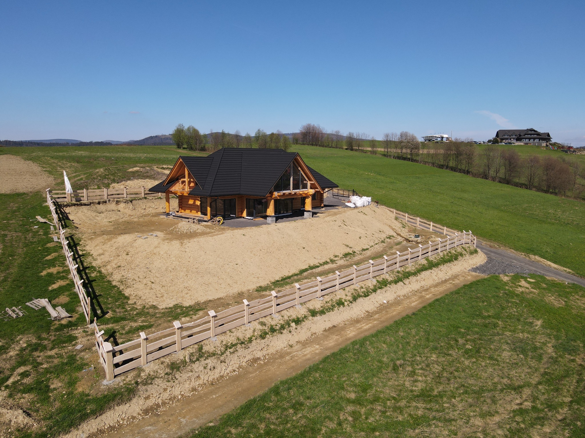 Widok z lotu ptaka na parterowy dom z bali z czarnym dachem, otoczony jasnym drewnianym płotem, na tle zielonego wzgórza i błękitnego nieba.