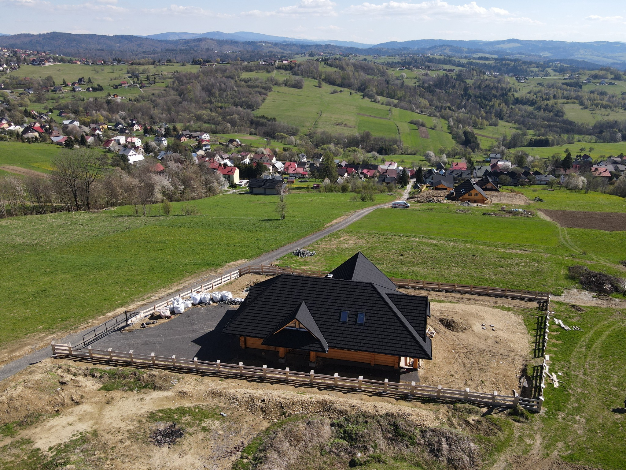 Nowoczesny, parterowy dom z bali z czarnym dachem, otoczony drewnianym płotem, widok z drona na tle pagórkowatego krajobrazu z zabudowaniami.