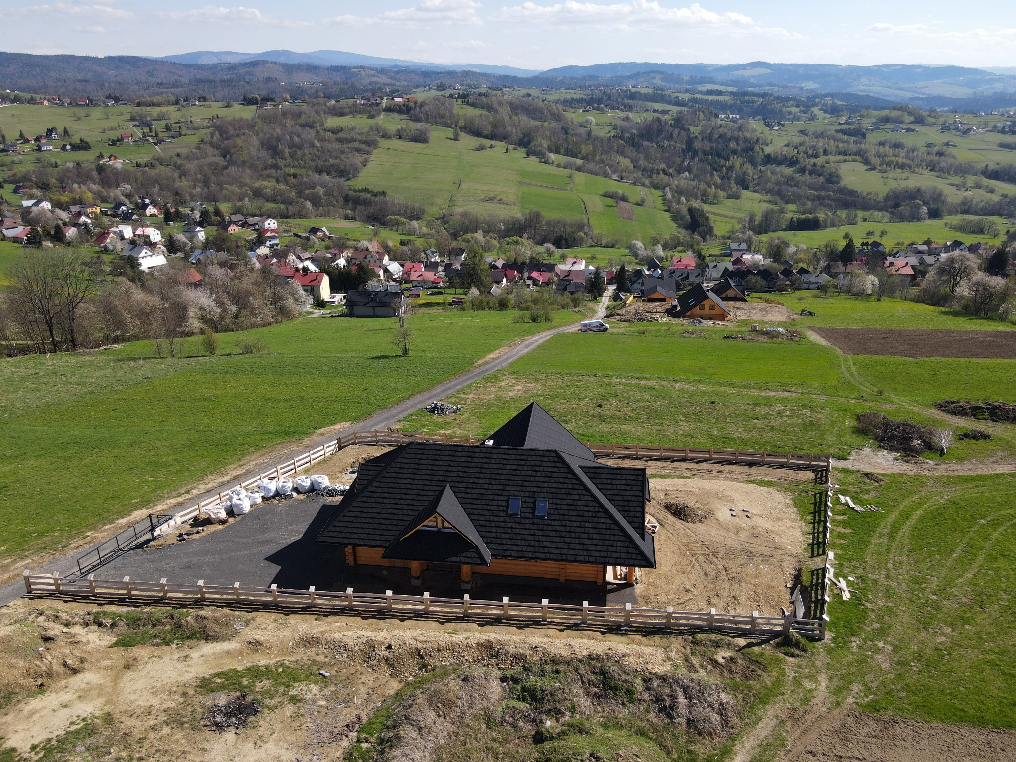 Nowo wybudowany, parterowy dom z bali z czarnym dachem, otoczony drewnianym płotem, na wzgórzu z widokiem na wieś i góry w tle, sfotografowany z lotu ptaka.