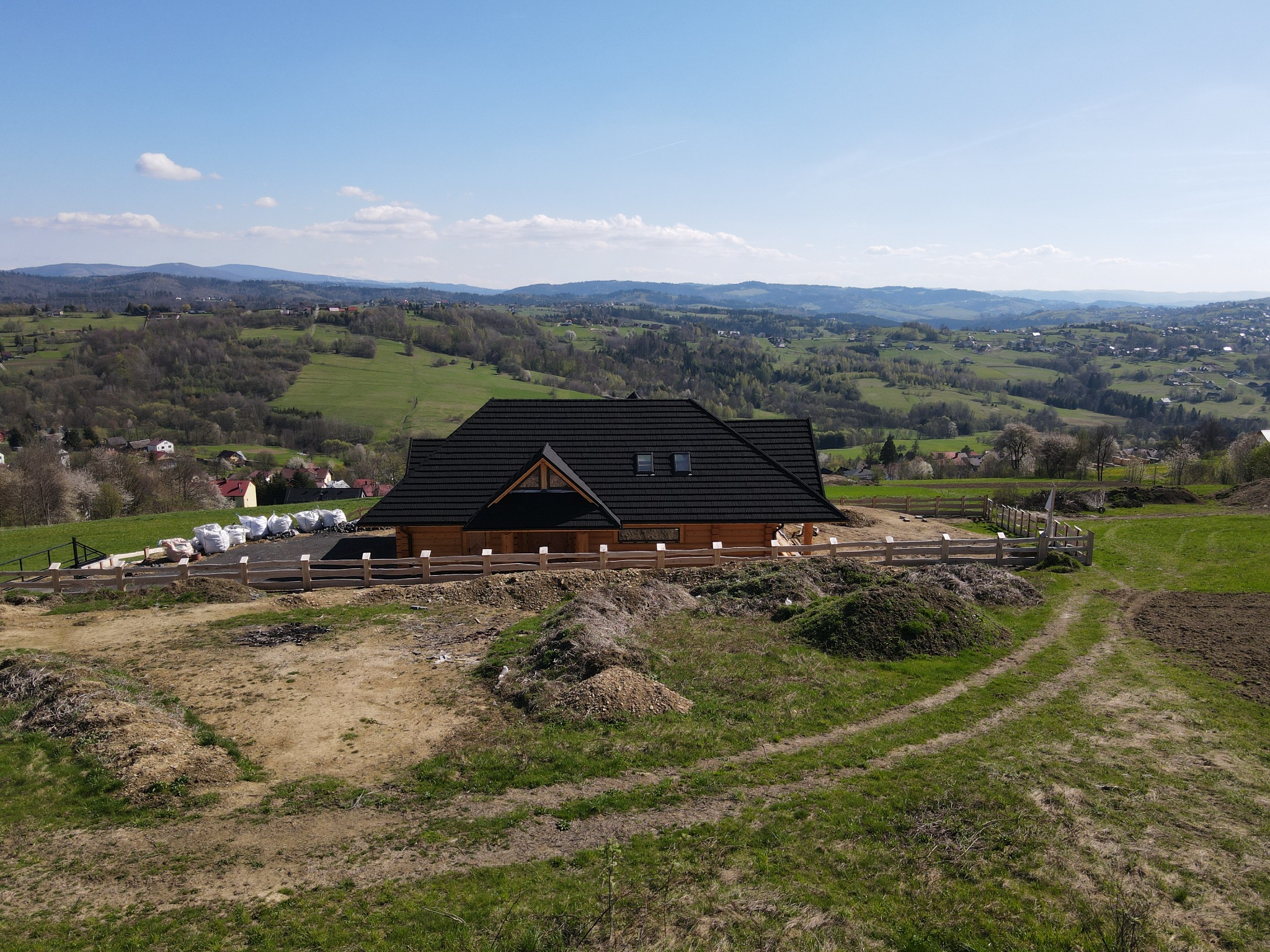 Surowy drewniany dom z bali z ciemnym dachem na wzgórzu, z widokiem na pagórkowaty krajobraz i błękitne niebo; teren wokół domu w trakcie zagospodarowania, widoczne worki z materiałami budowlanymi.