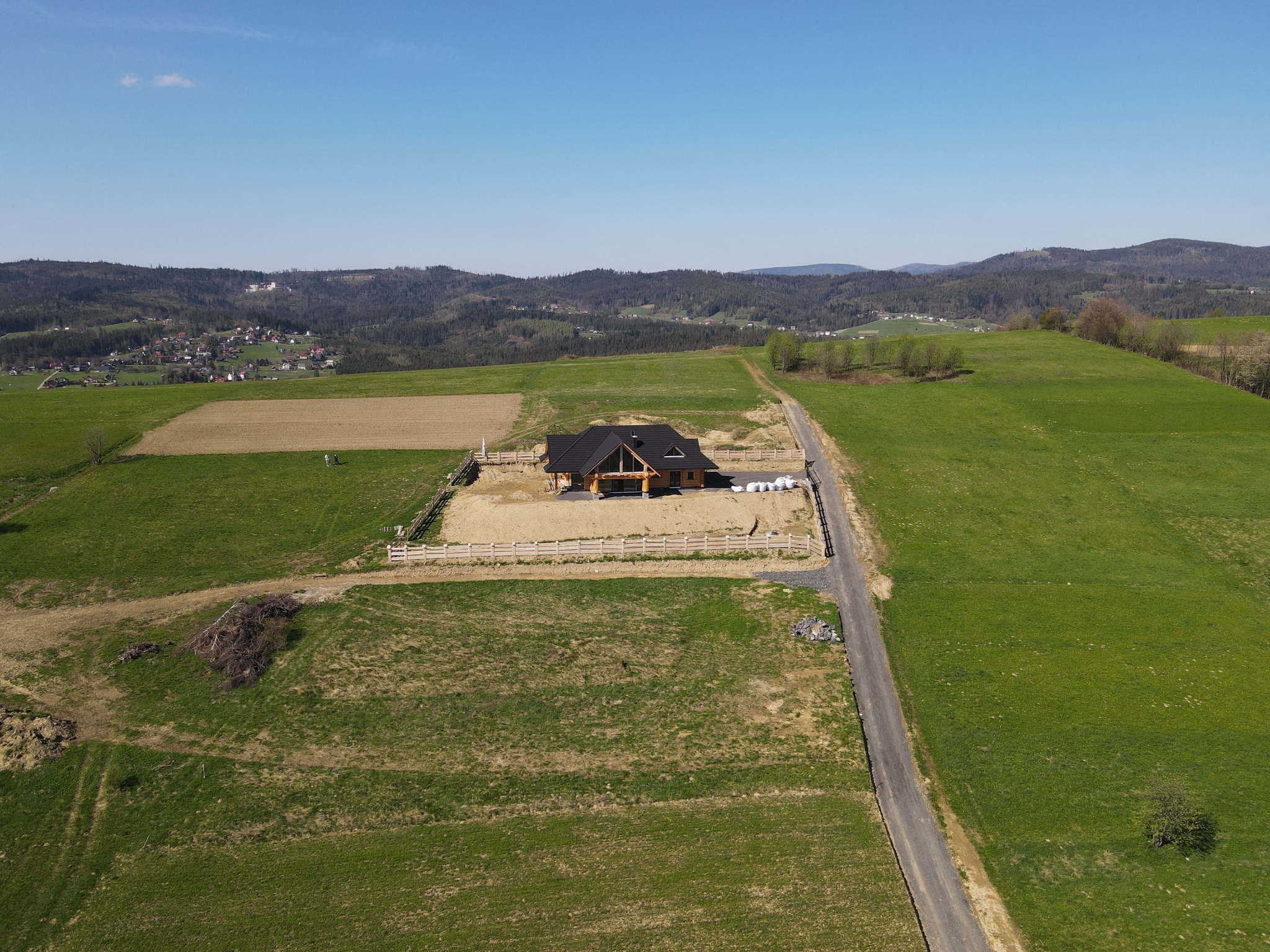 Szeroki kadr z lotu ptaka na budowę domu z bali z czarnym dachem na rozległym, zielonym terenie wiejskim, z widocznymi górami w tle i drogą dojazdową.