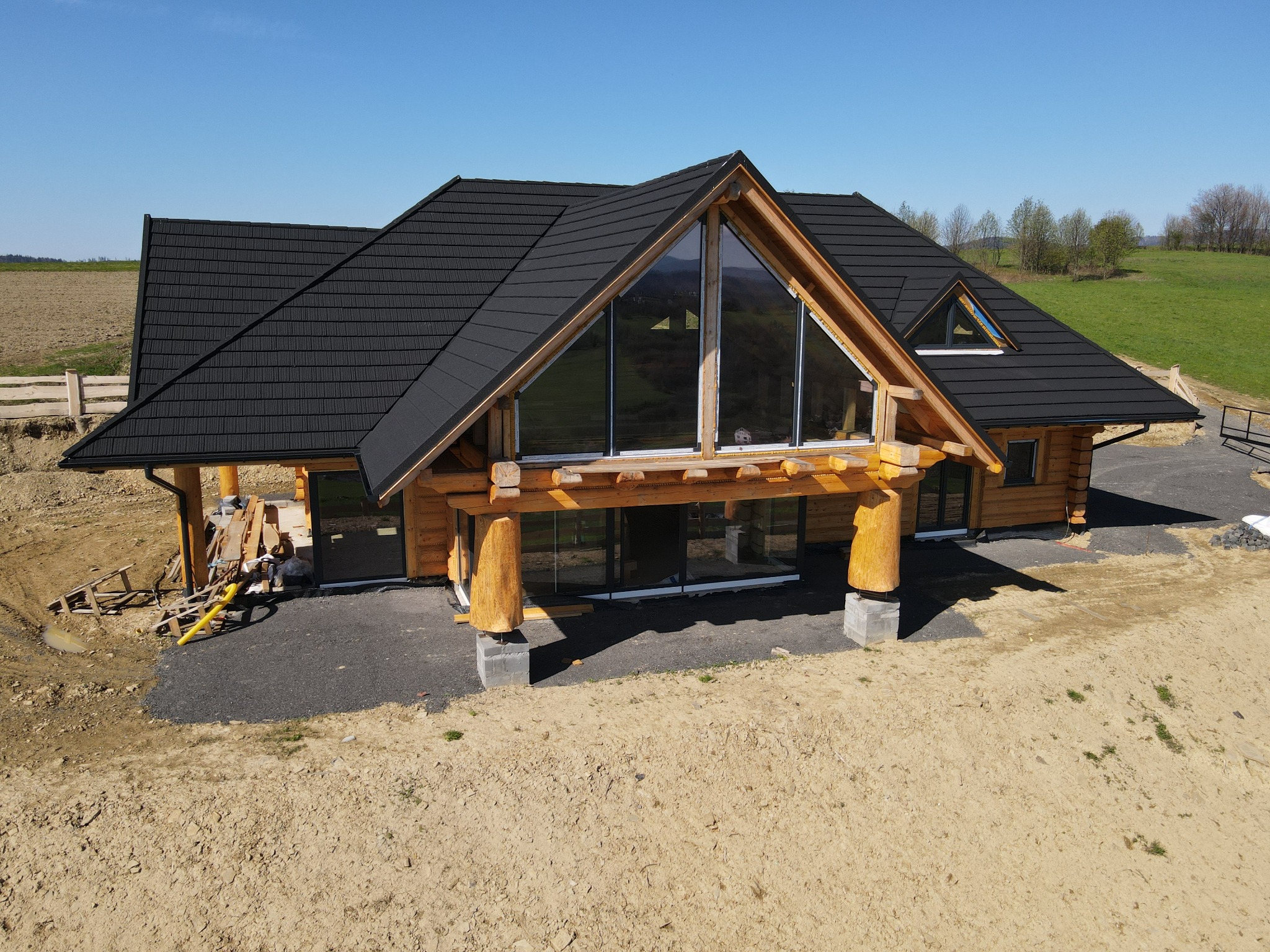 Surowy stan budowy domu z bali z czarnym dachem, dużym oknem szczytowym i drewnianymi kolumnami na betonowych podstawach, widok z góry na plac budowy.