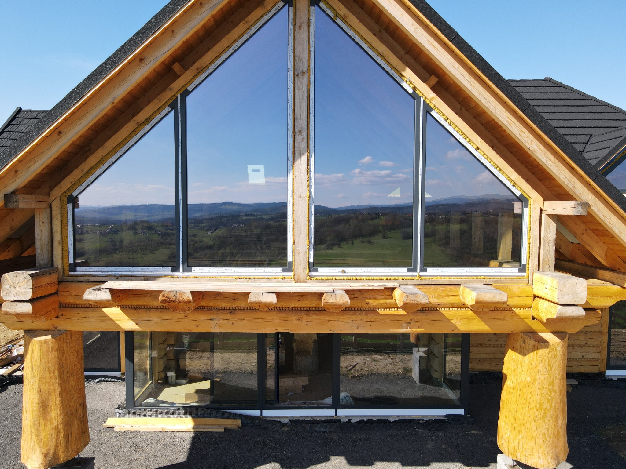 Fragment drewnianego domu w trakcie budowy, widoczne okna z czarnymi ramami, konstrukcja z bali, tło z zielonymi wzgórzami i błękitnym niebem.
