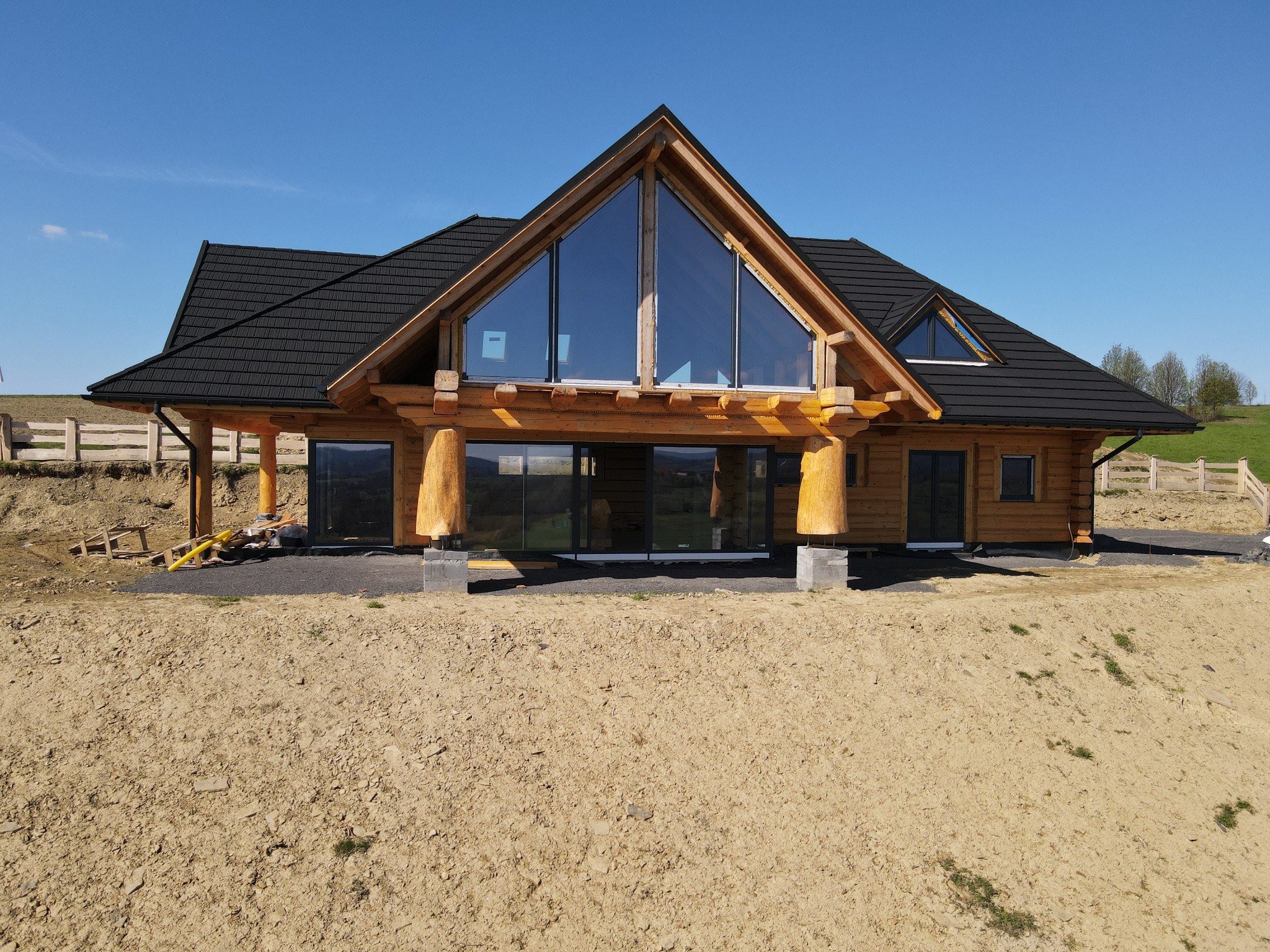 Surowy stan budowy domu z bali z dużym trójkątnym oknem szczytowym, widoczne tymczasowe podpory i niezagospodarowany teren wokół, czarna dachówka.