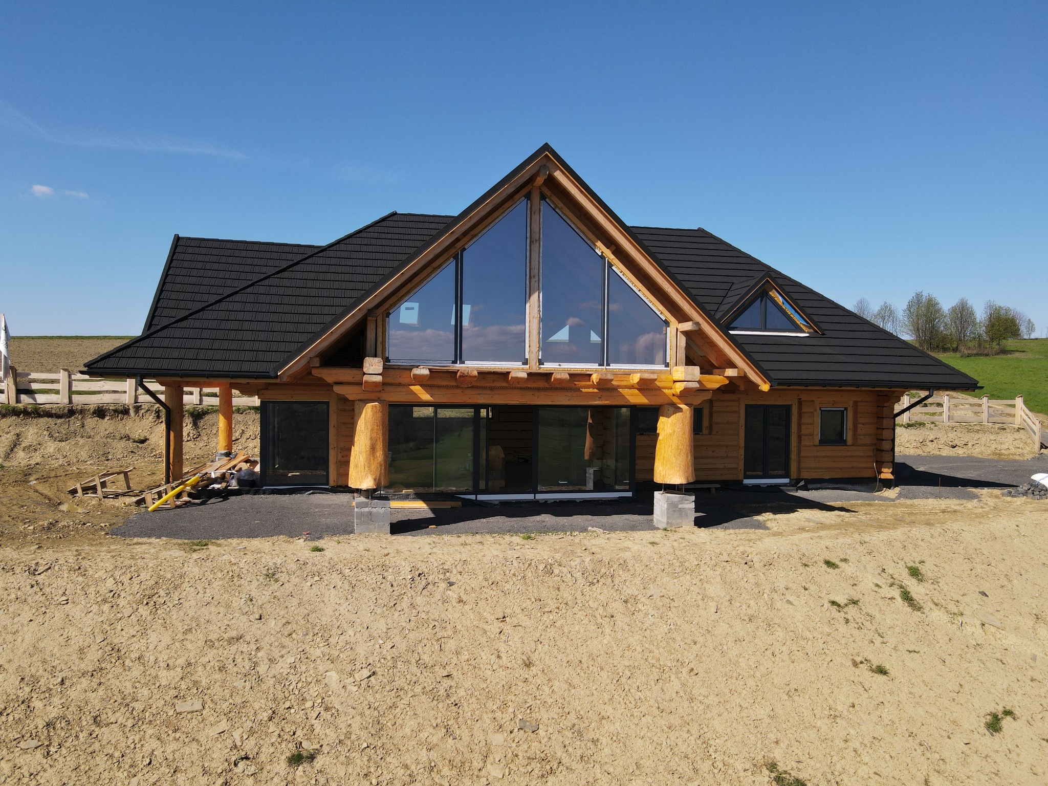 Nowoczesny dom z bali w trakcie budowy, widoczne surowe drewno, duże przeszklenia i czarna dachówka, teren wokół jeszcze niezagospodarowany.