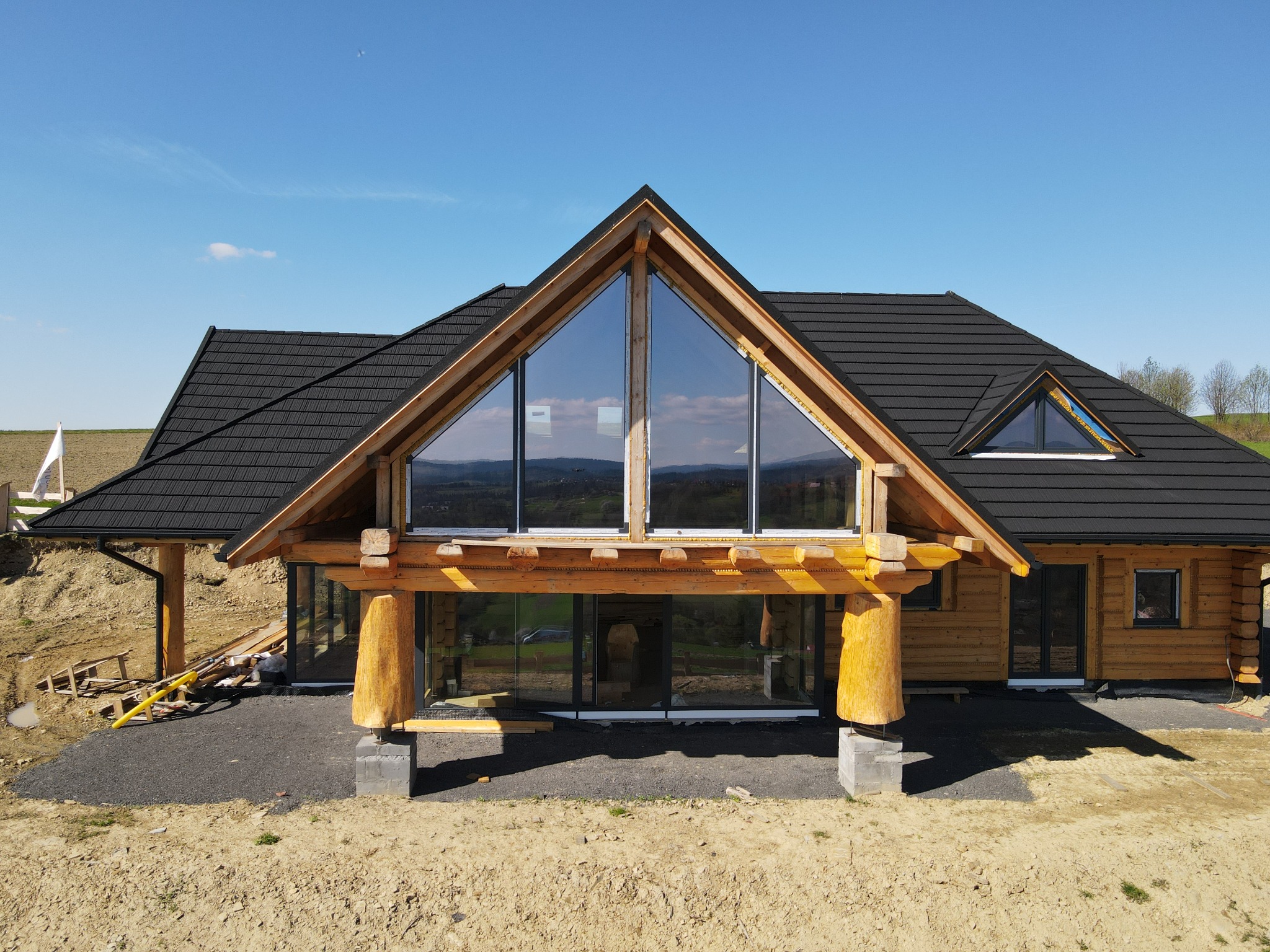 Surowy stan budowy domu z bali w Istebnej, z charakterystycznym trójkątnym oknem szczytowym i czarną dachówką, osadzony na betonowych fundamentach z widocznymi elementami konstrukcyjnymi.