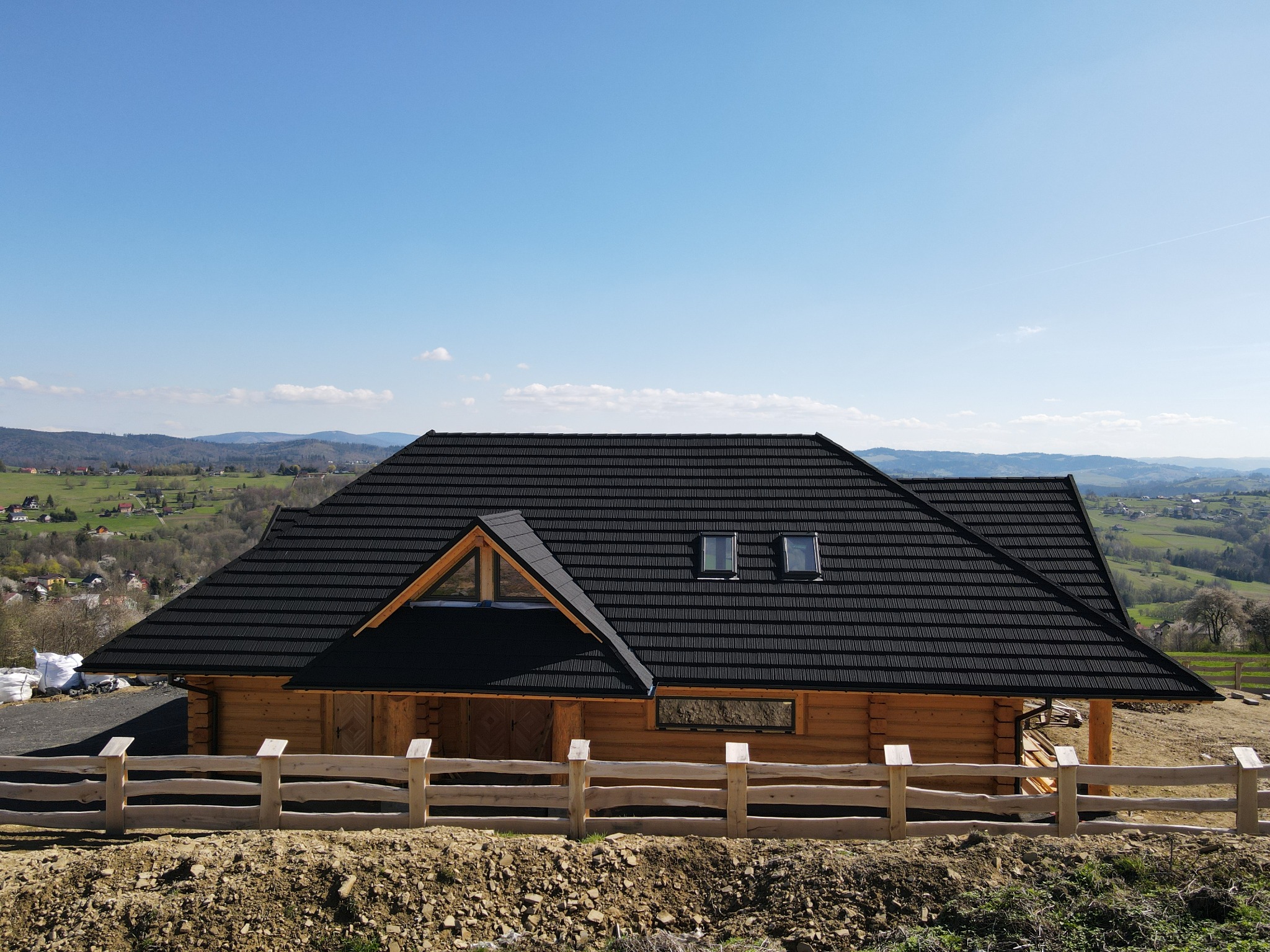 Nowoczesny dom z bali z czarnym dachem, widok z zewnątrz na tle pagórkowatego krajobrazu i błękitnego nieba, z surowym drewnianym ogrodzeniem w pierwszym planie.