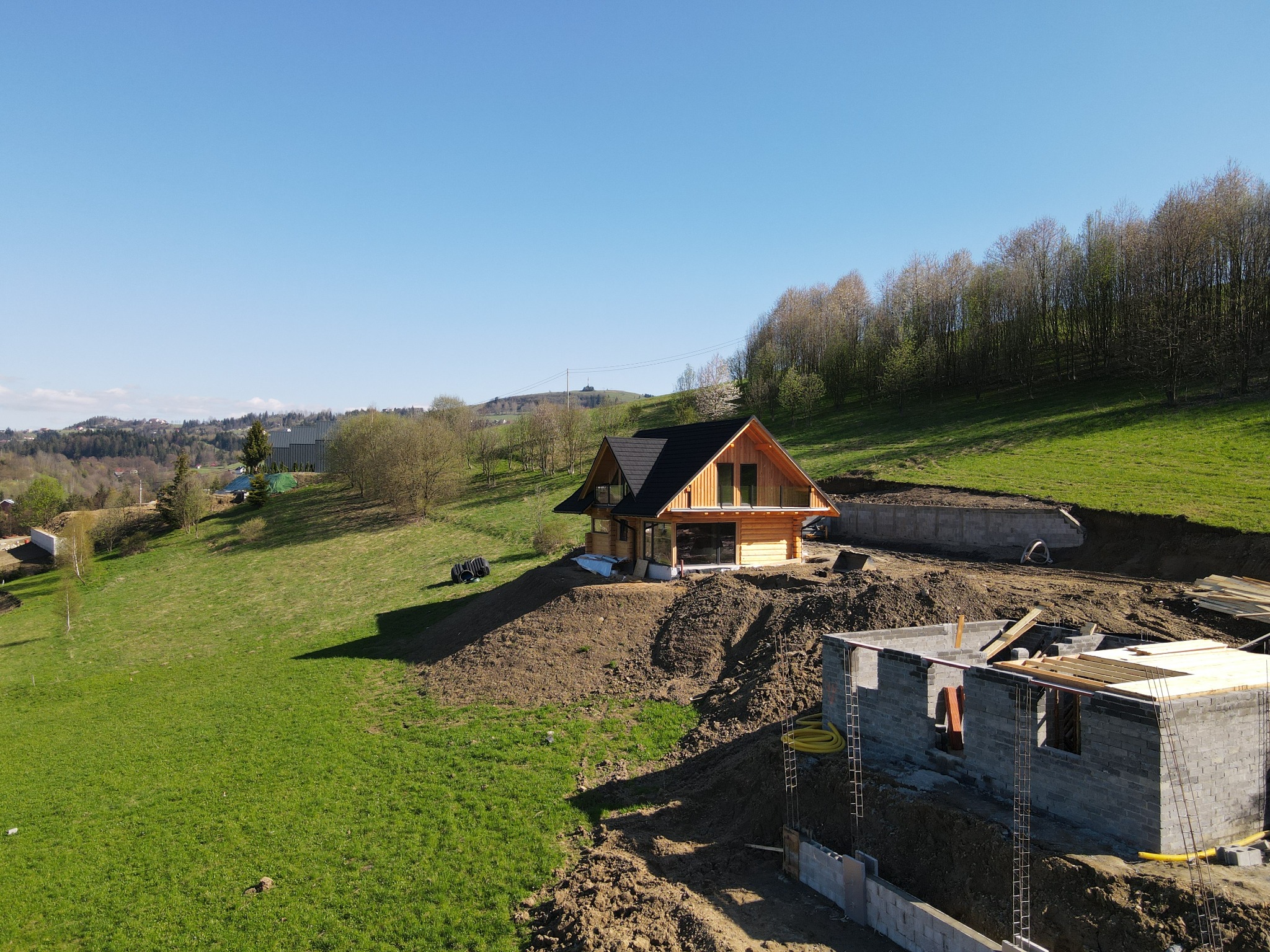 Wznoszony dom z bali z ciemnym dachem na wzgórzu, widok z góry, obok fundamenty drugiego budynku, w tle zielone łąki i las.