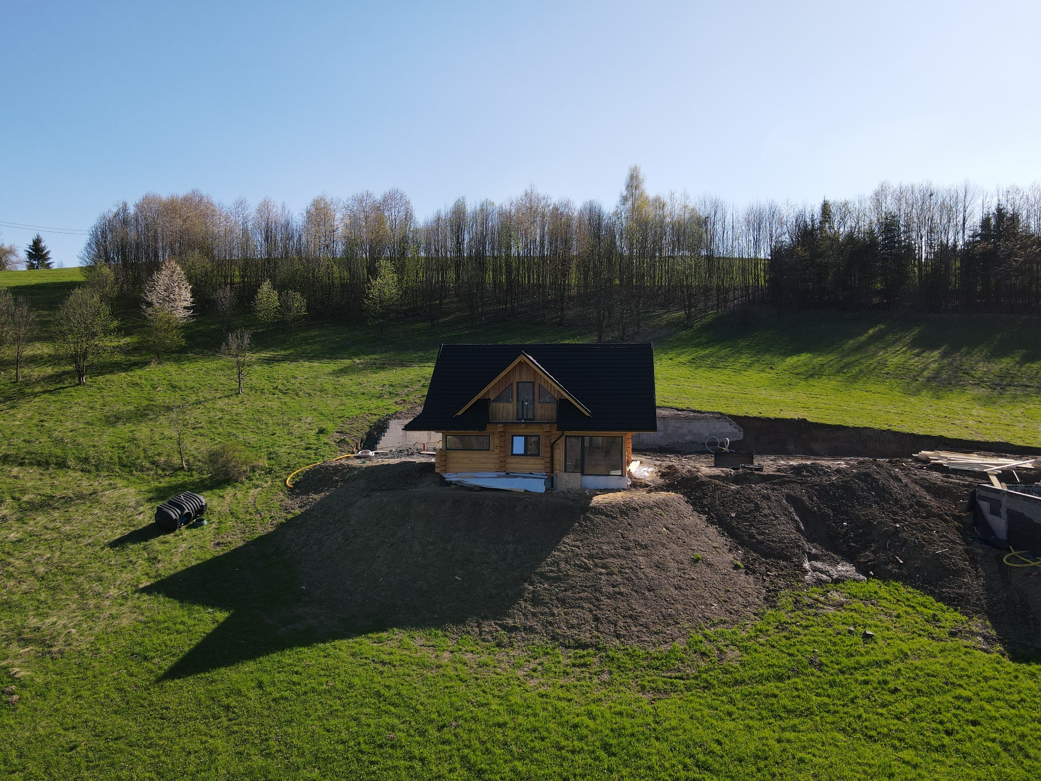 Nowoczesny, drewniany dom z ciemnym dachem w trakcie budowy na wzgórzu, widok z lotu ptaka, w tle drzewa i błękitne niebo.