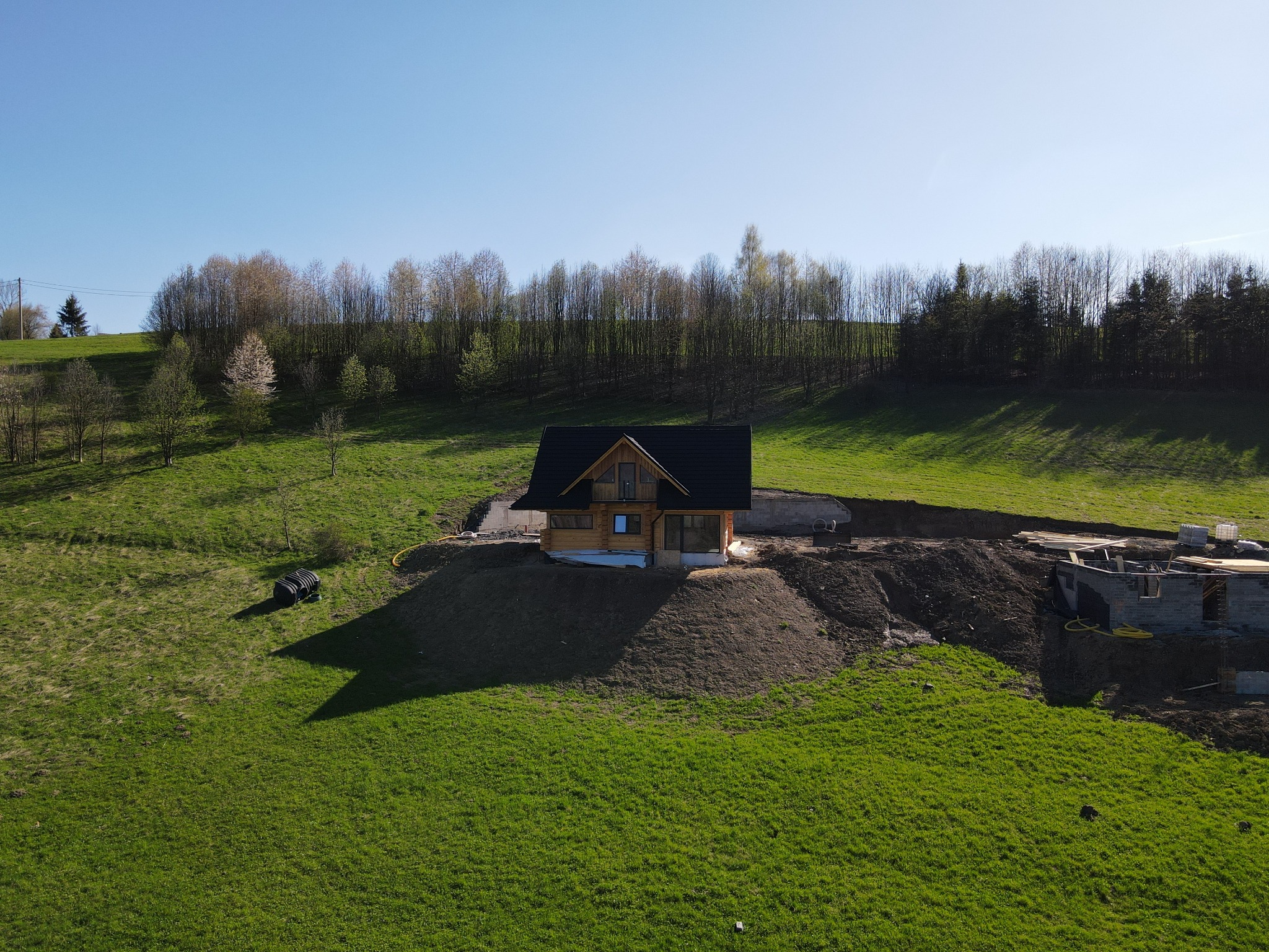 Widok z lotu ptaka na drewniany dom w trakcie budowy, z widocznym fundamentem i elementami konstrukcyjnymi, otoczony zielonym trawnikiem i drzewami na wzgórzu, przy słonecznej pogodzie.