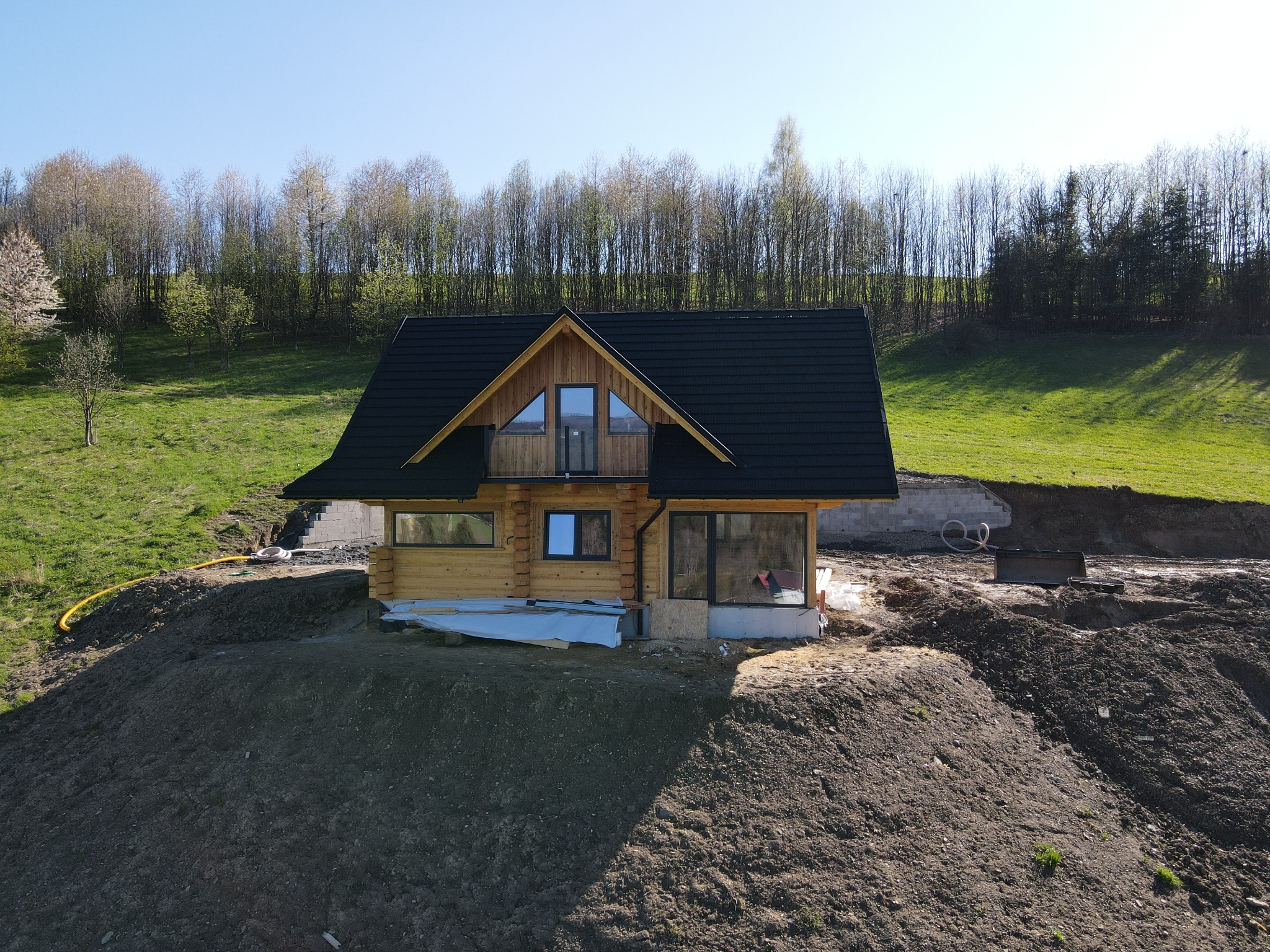 Surowy, drewniany dom z bali z czarnym dachem w trakcie budowy na wzgórzu z odsłoniętą ziemią, z widocznymi drzewami w tle.