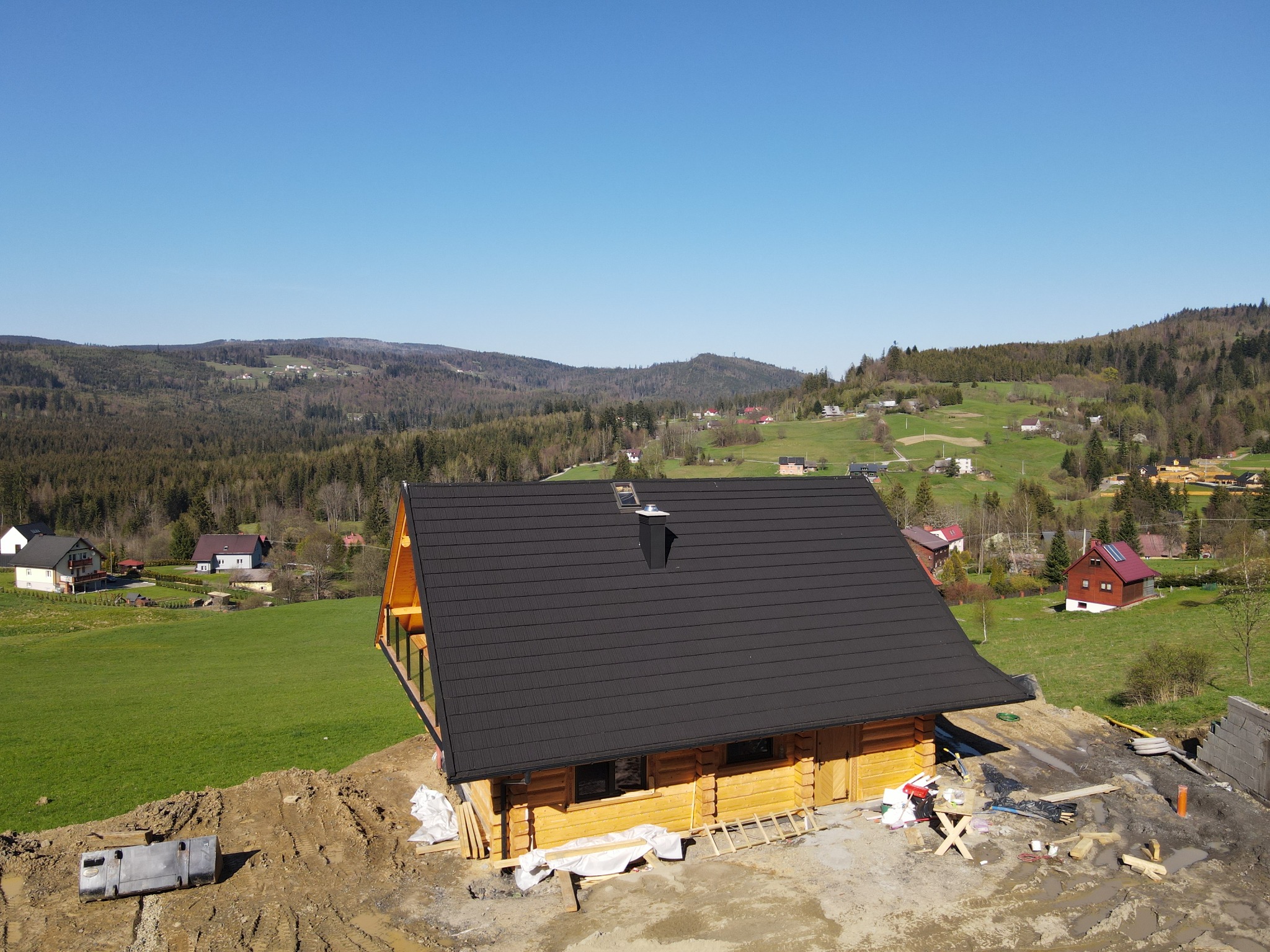 Nowo wybudowany drewniany dom z bali z czarnym dachem na tle pagórkowatego krajobrazu, widok z góry na plac budowy z widocznymi materiałami i narzędziami.