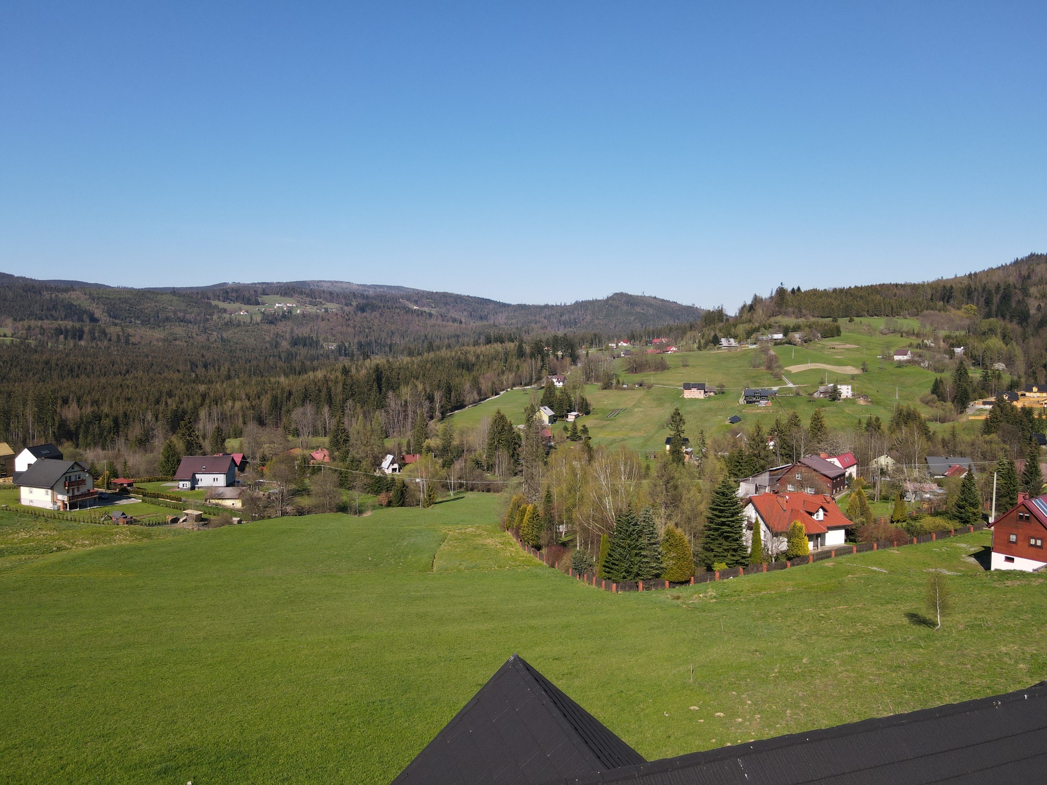 Panoramiczny widok na wiejski krajobraz z zabudowaniami, lasami i wzgórzami, widziany z dachu pokrytego czarną dachówką.