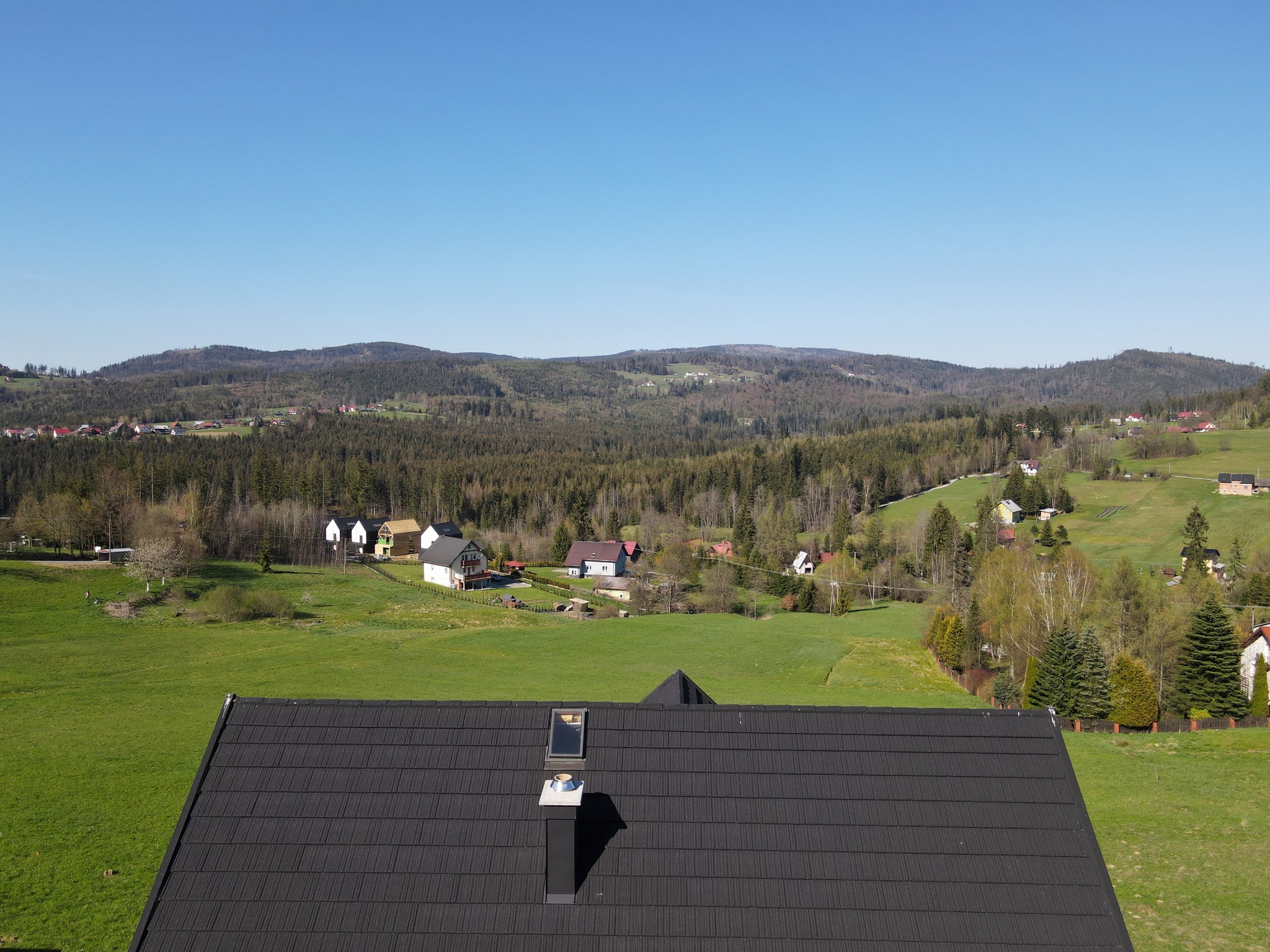 Widok z góry na nowy, czarny dach z blachodachówki z kominem i oknem dachowym na tle zielonego krajobrazu z domami i lasami.