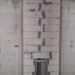 Wewnętrzna ściana z betonu komórkowego w trakcie budowy komina wentylacyjnego, widoczne przewody elektryczne w peszlach, puszka instalacyjna podtynkowa z pomarańczowym deklem i wystające kable.