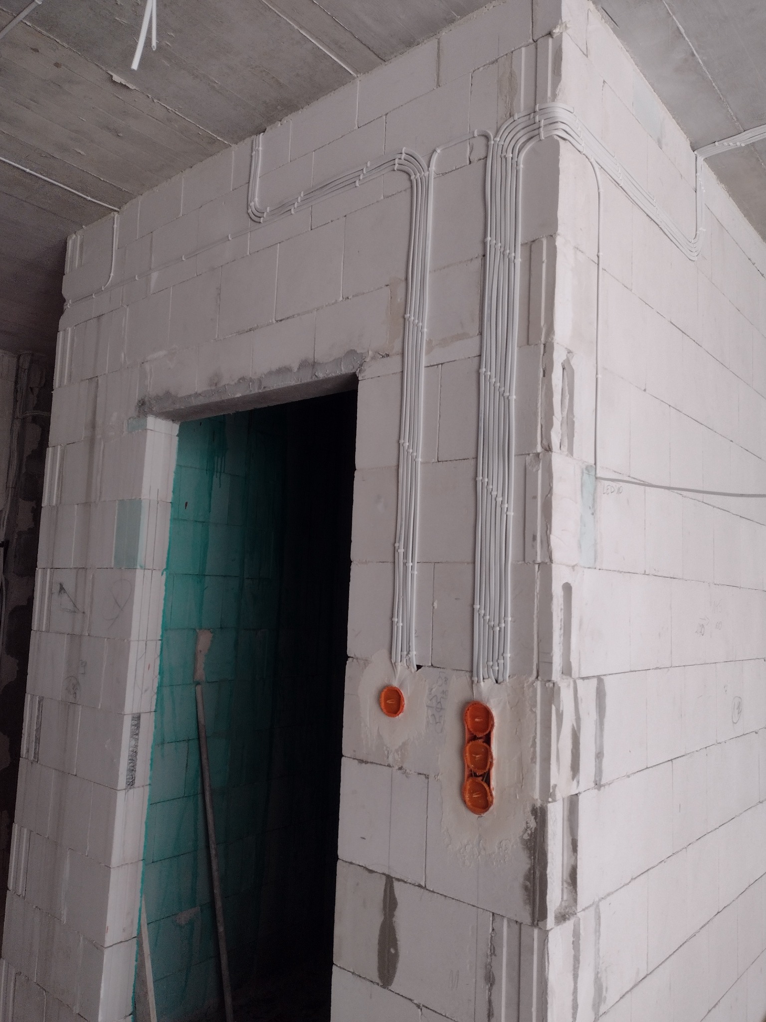 Wewnętrzne okablowanie elektryczne w ścianie z białych bloczków, widoczne rury instalacyjne pionowo ułożone wzdłuż narożnika, pomarańczowe puszki pod gniazdka, surowy stan wykończenia.