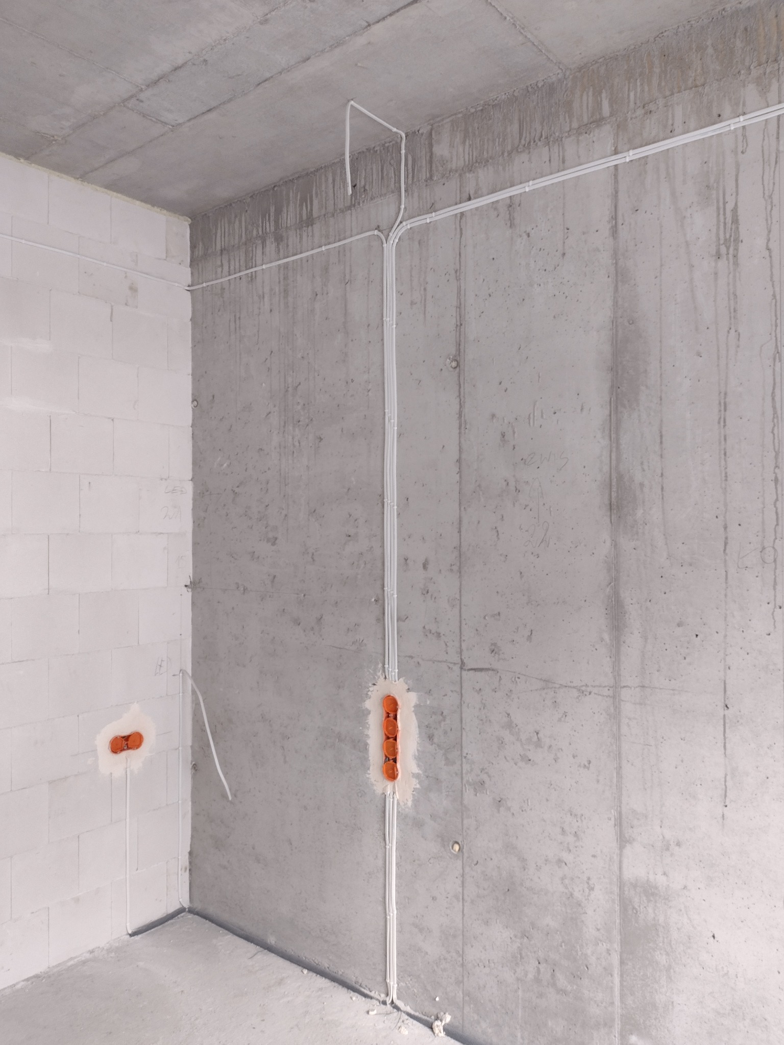 Surowy stan wykończenia wnętrza: widoczne kable elektryczne prowadzone po ścianie z betonu architektonicznego i ścianie z białego bloczka, puszki instalacyjne w pomarańczowym kolorze.