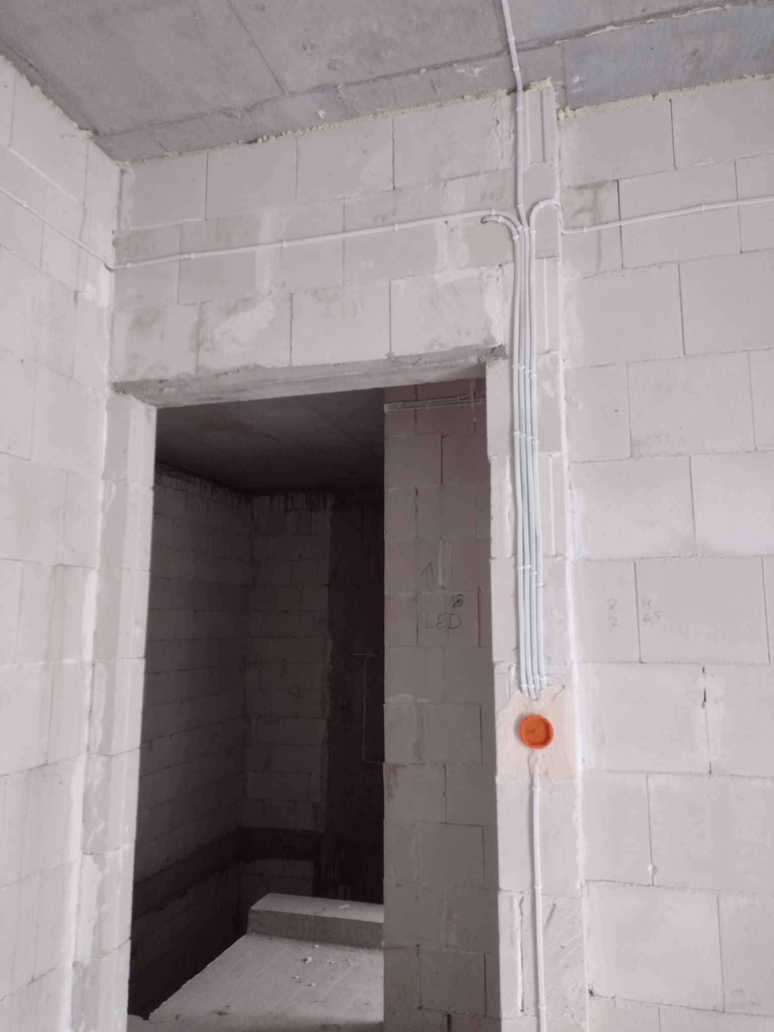 Wewnętrzna ściana z betonu komórkowego w stanie surowym z poprowadzonymi przewodami elektrycznymi w białych rurkach, widoczna pomarańczowa puszka instalacyjna i otwór drzwiowy prowadzący...