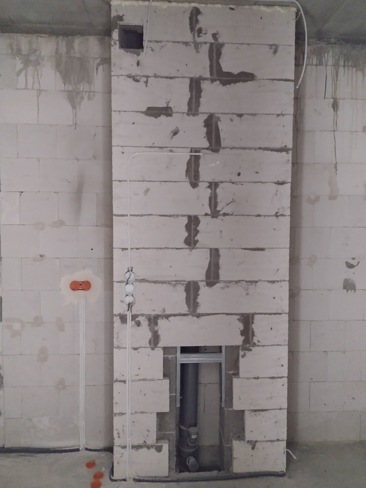 Wewnętrzna ściana z betonu komórkowego w trakcie budowy komina wentylacyjnego, widoczne przewody elektryczne w peszlach, puszka instalacyjna podtynkowa z pomarańczowym deklem i wystające kable.