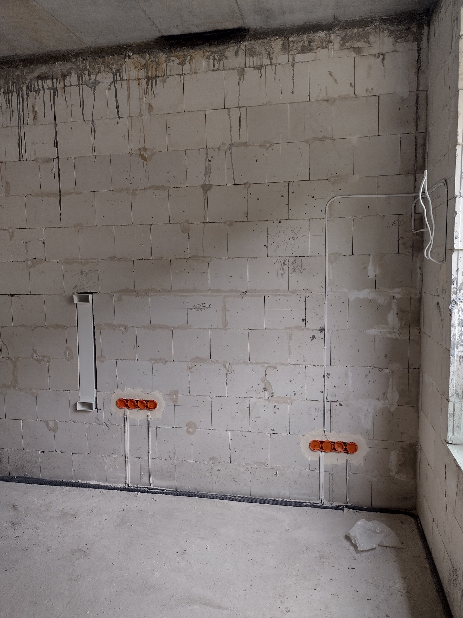 Surowa ściana z betonu komórkowego z widocznymi przewodami elektrycznymi w peszlach i puszkami pod gniazdka, widoczne ślady po zalaniu na suficie, w tle fragment okna.