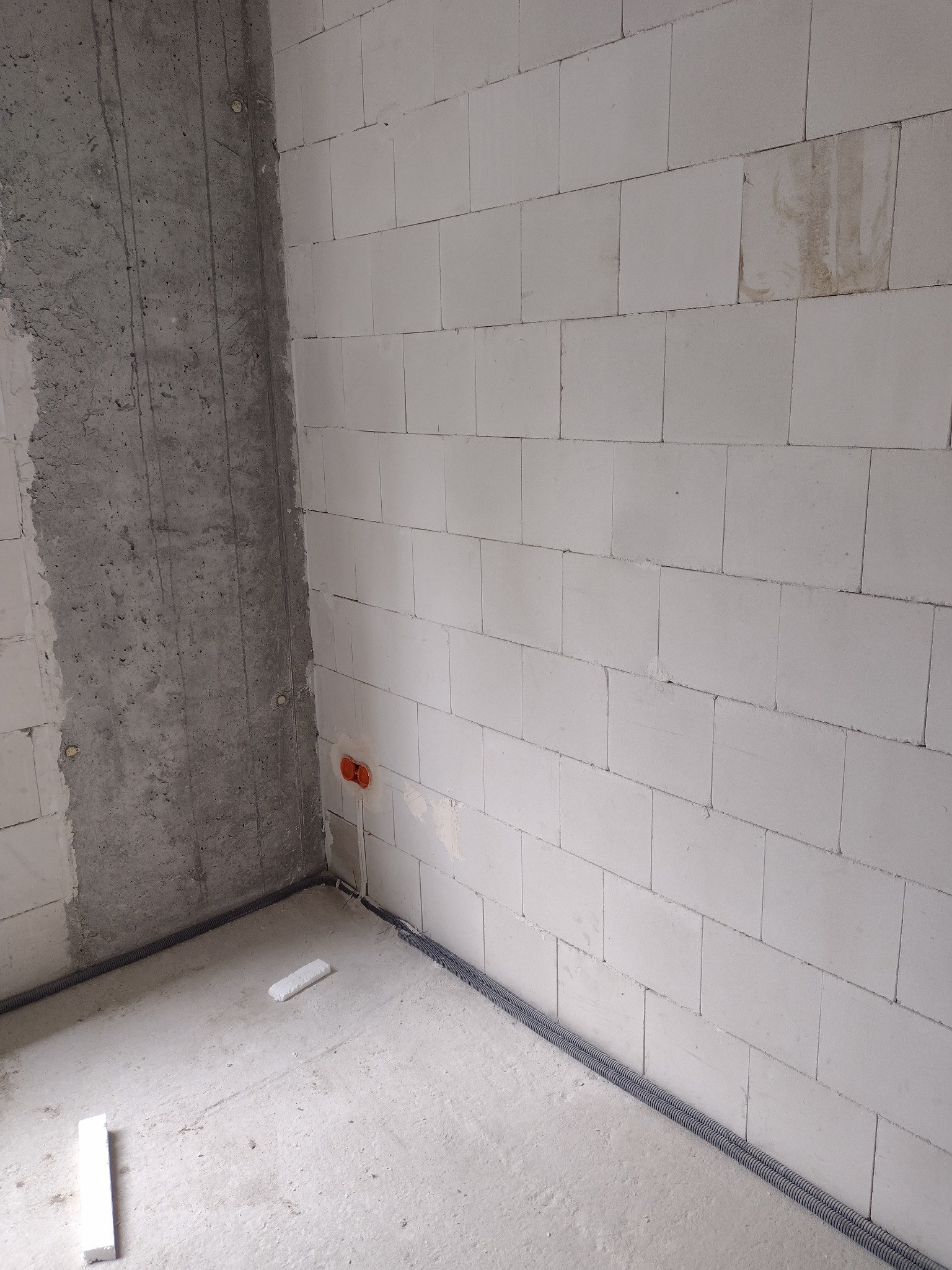 Wewnętrzny róg surowego pomieszczenia z instalacją elektryczną w peszlach przy podłodze, widoczne puszki instalacyjne w ścianie z betonu komórkowego i fragment surowej ściany betonowej.