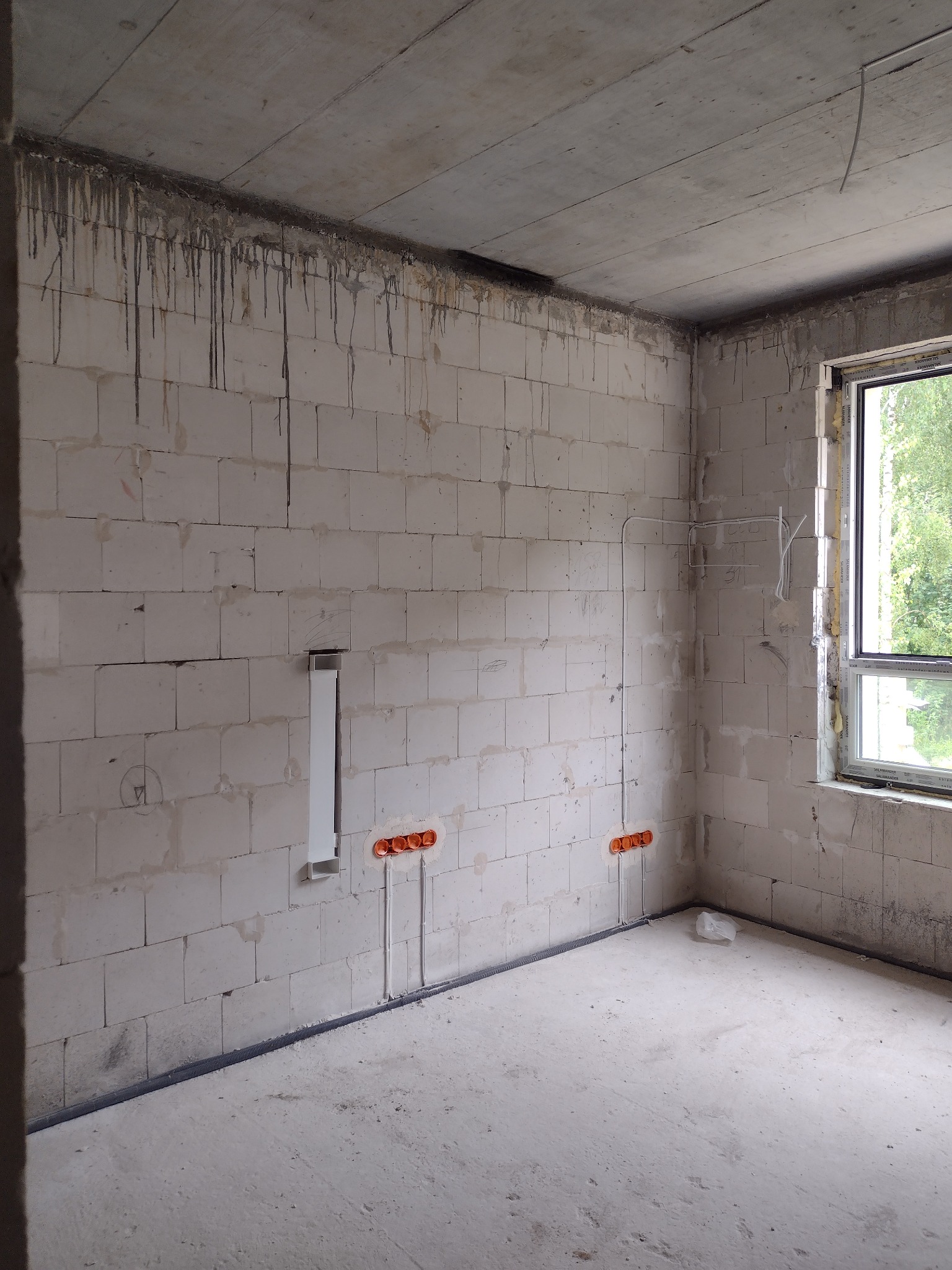 Surowy pokój w stanie deweloperskim z widocznymi przewodami elektrycznymi w ścianie z betonu komórkowego oraz puszkami instalacyjnymi, okno z widokiem na zieleń, sufit betonowy z zaciekami.