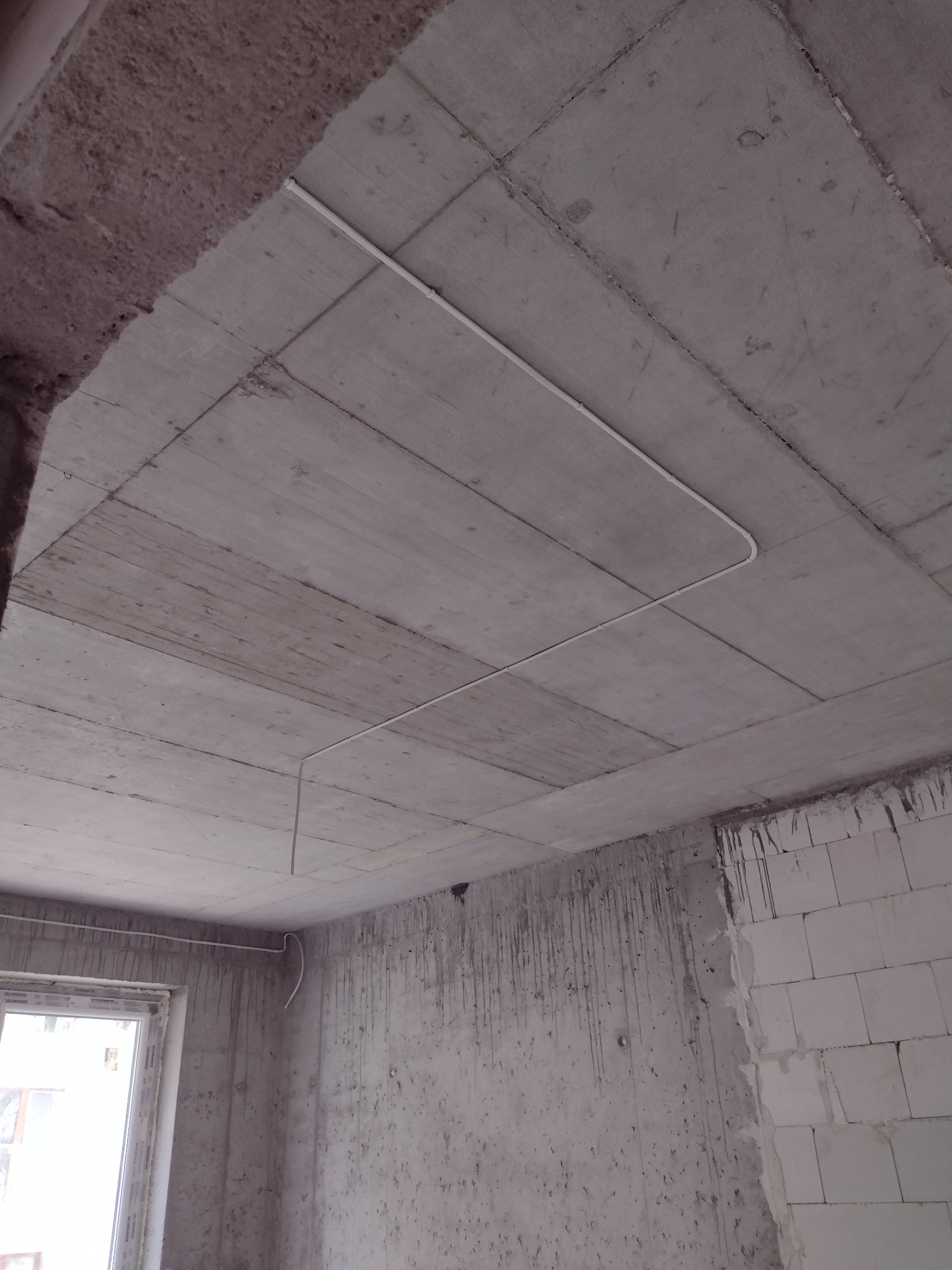 Szara surowa betonowa płyta stropowa z poprowadzonymi białymi rurkami instalacyjnymi, widok z dołu w surowym wnętrzu budynku w trakcie budowy.