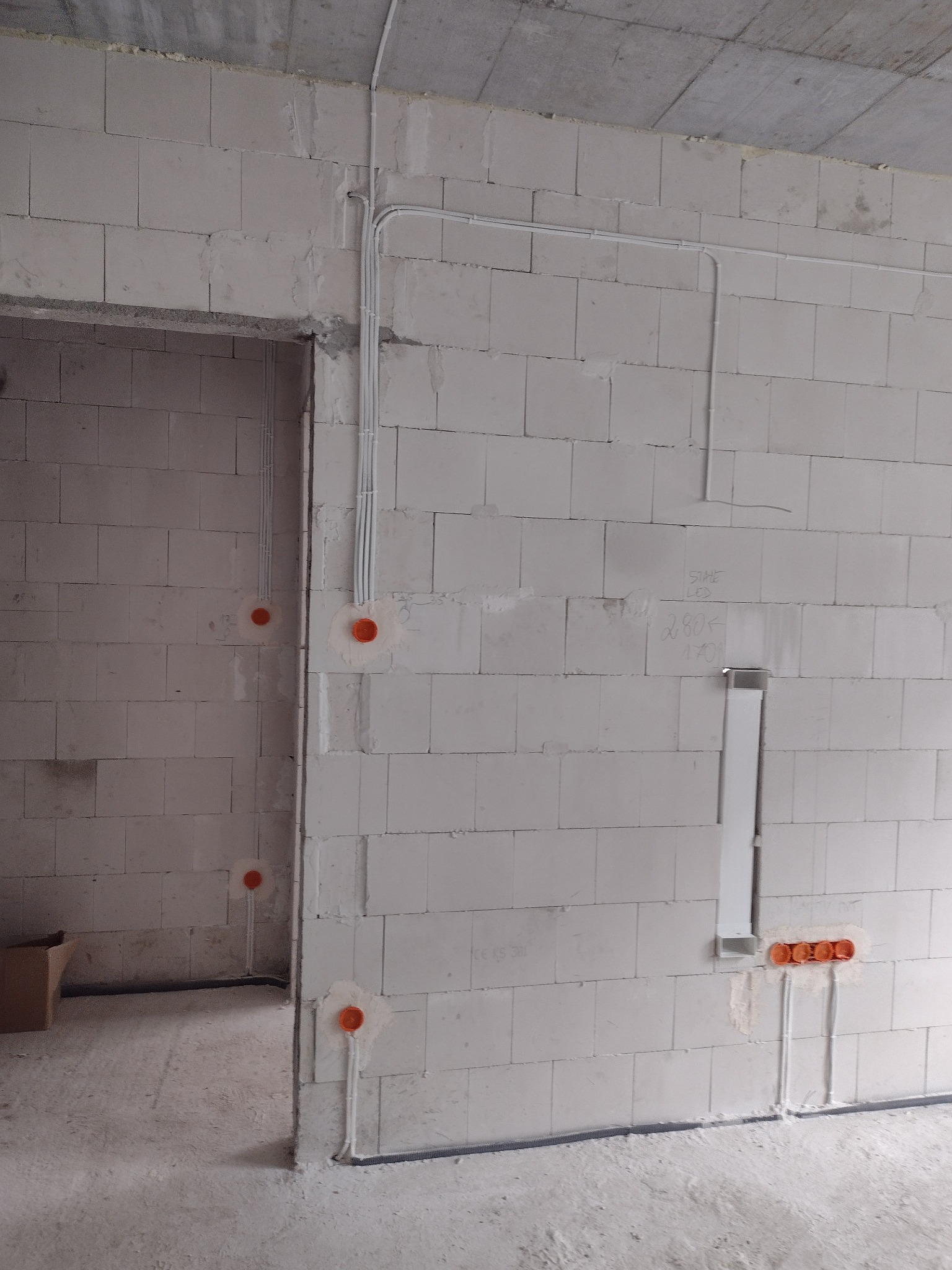 Surowy stan instalacji elektrycznej w nowym budynku: białe kable w peszlach prowadzone po ścianach z bloczków, pomarańczowe puszki instalacyjne, widoczne napisy ołówkiem na ścianie.