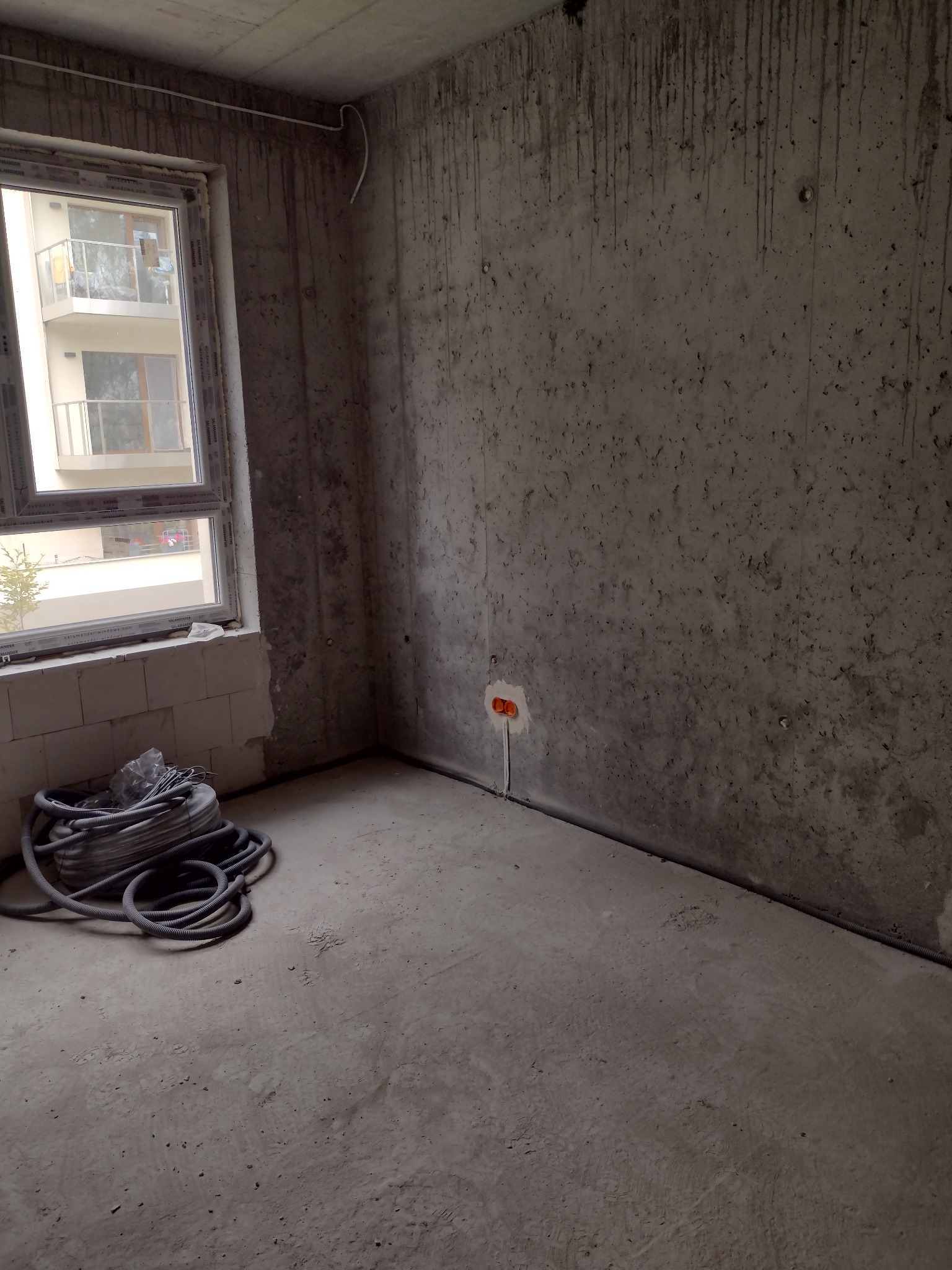 Surowe wnętrze w trakcie prac wykończeniowych: widoczne kable elektryczne wystające ze ściany, szpula kabli na podłodze, betonowe ściany z zaciekami i nowe okno z widokiem na balkon sąsiedniego...