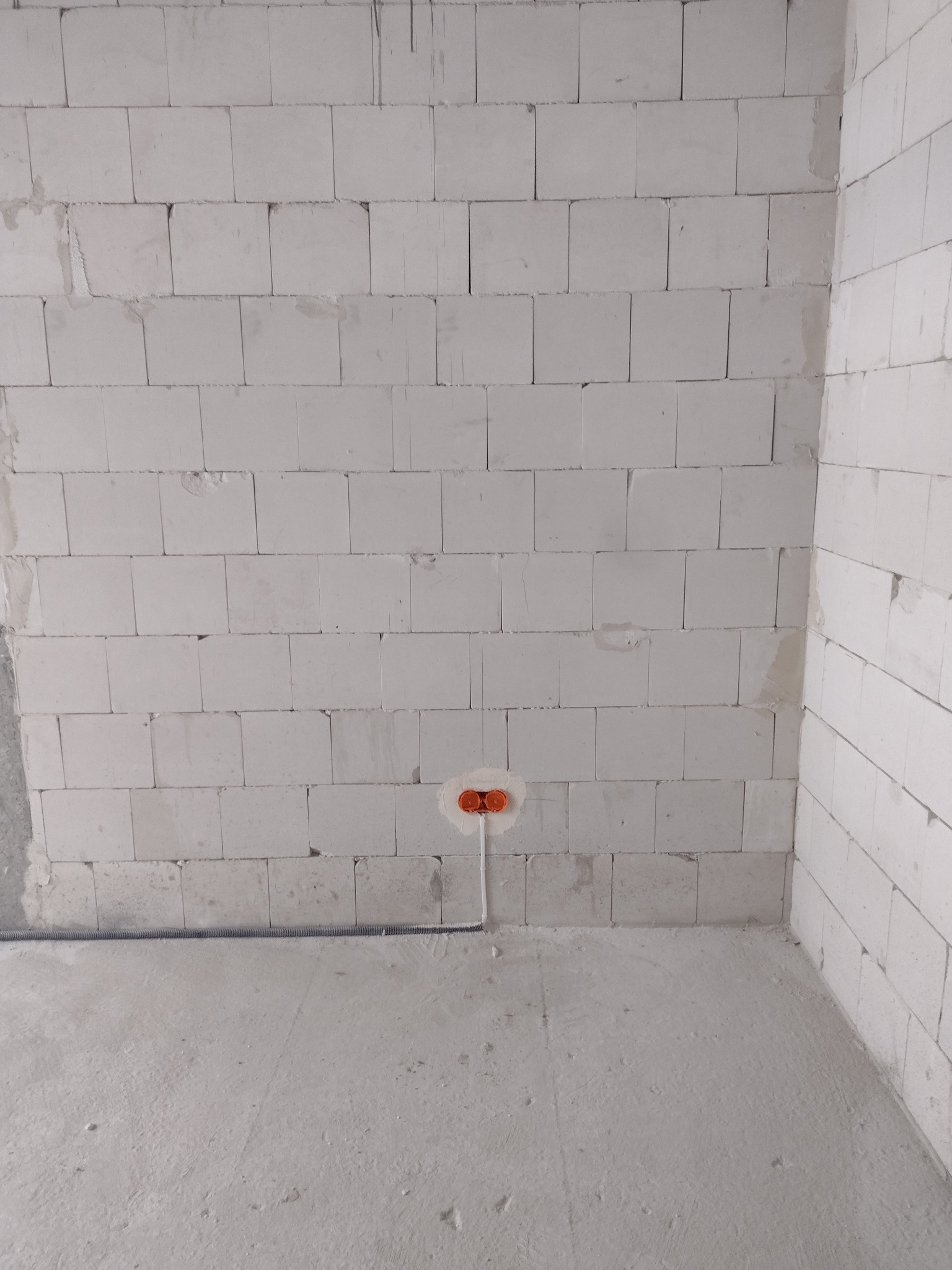 Surowa ściana z betonu komórkowego z zamontowaną, pomarańczową puszką elektryczną i wystającymi przewodami, widok z dołu.