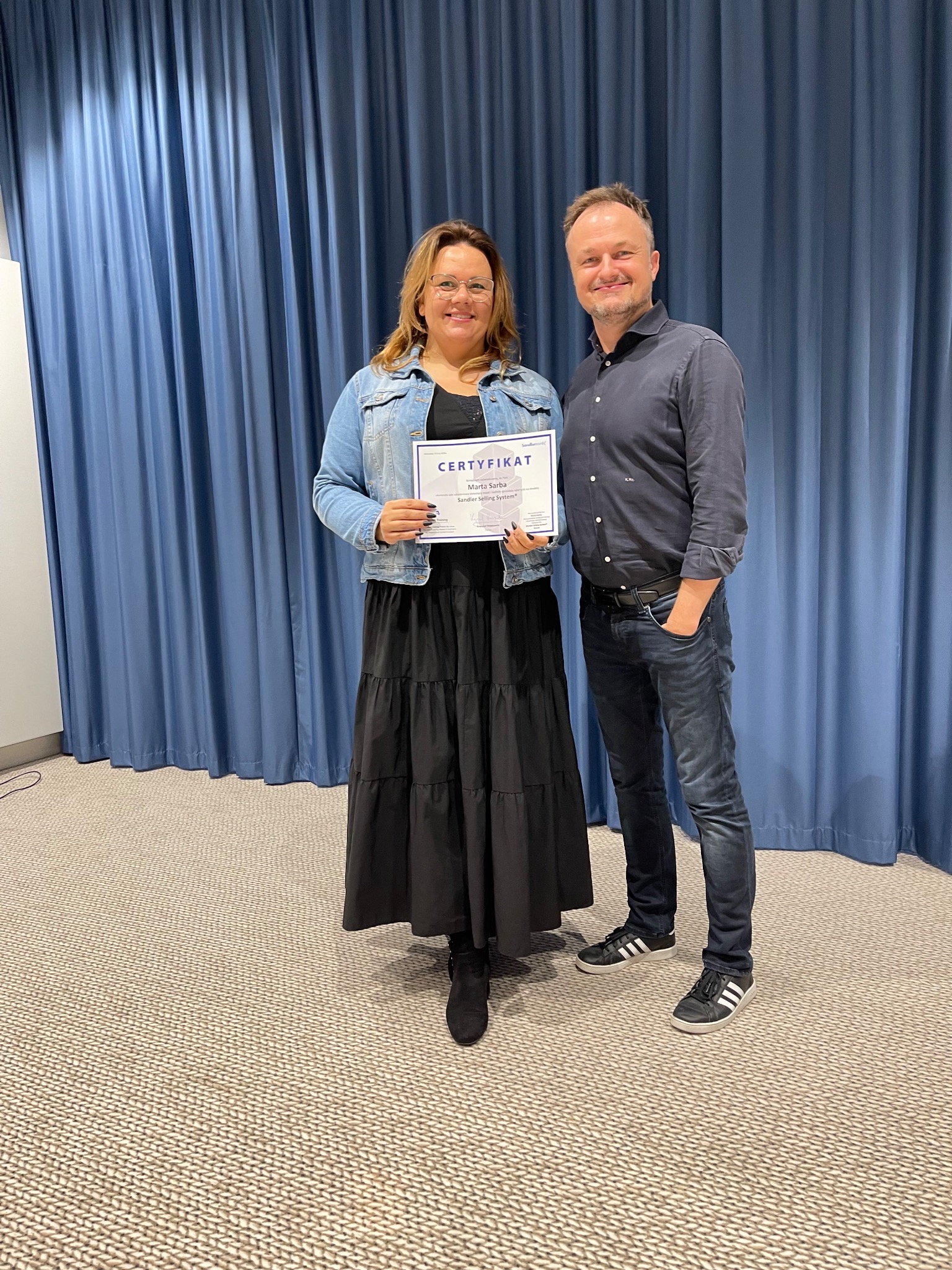 Kobieta w dżinsowej kurtce i czarnej sukience trzyma certyfikat, stojąc obok mężczyzny na tle niebieskich zasłon.