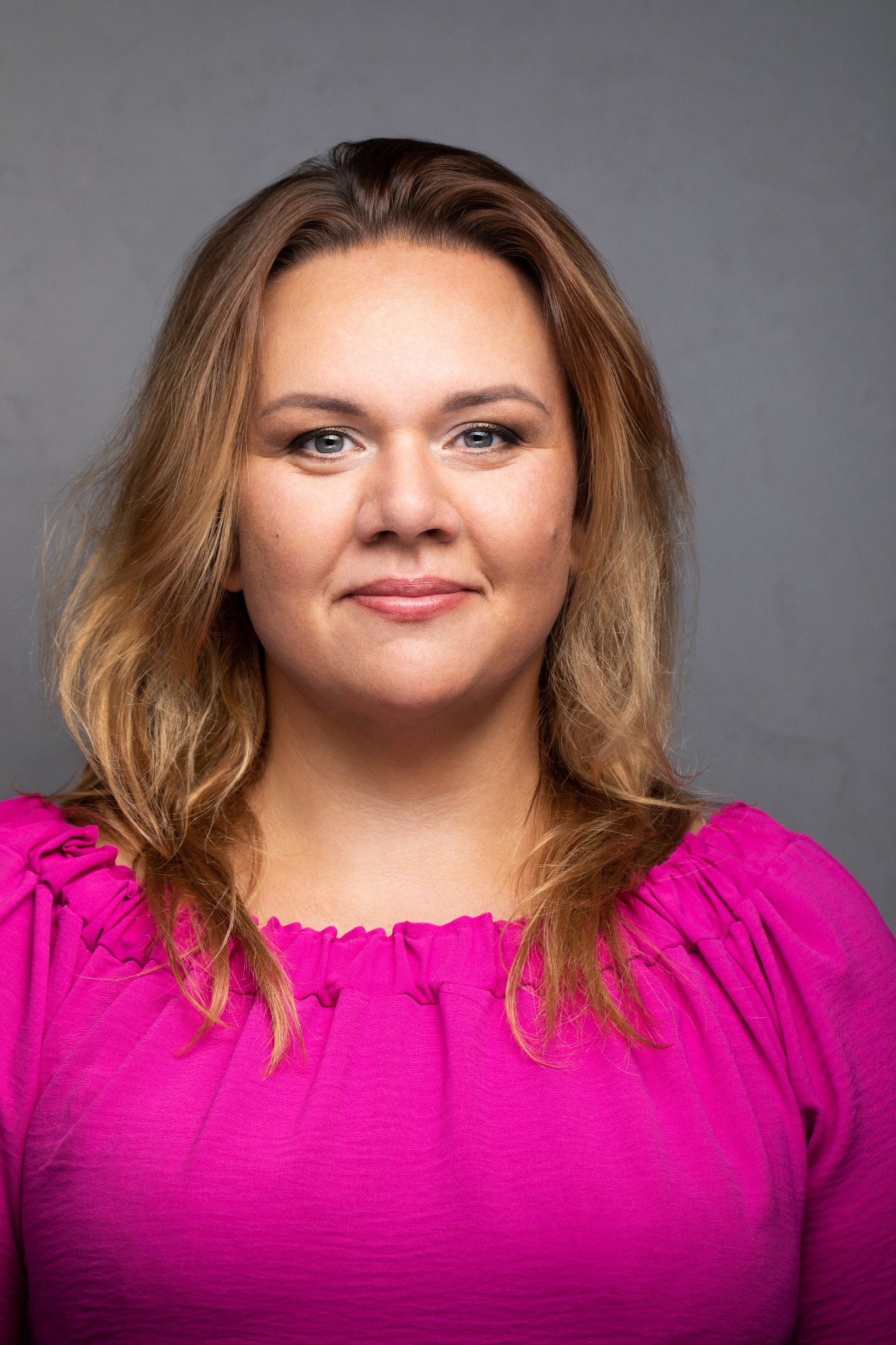 Portret uśmiechniętej kobiety z blond włosami w różowej bluzce na szarym tle.