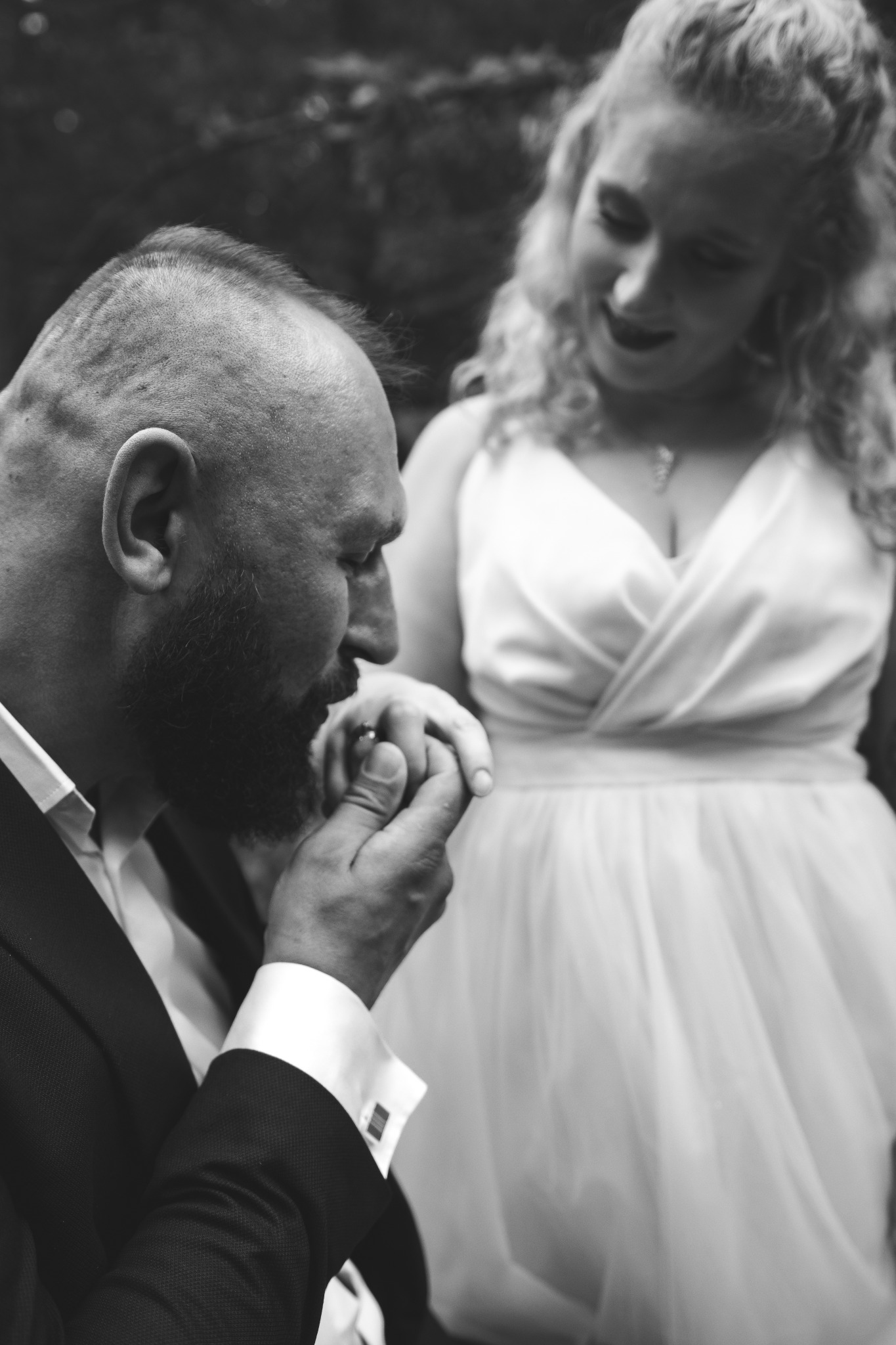 Czarno-białe zbliżenie: mężczyzna w garniturze całuje dłoń uśmiechniętej kobiety w sukni ślubnej, romantyczna scena z uroczystości.