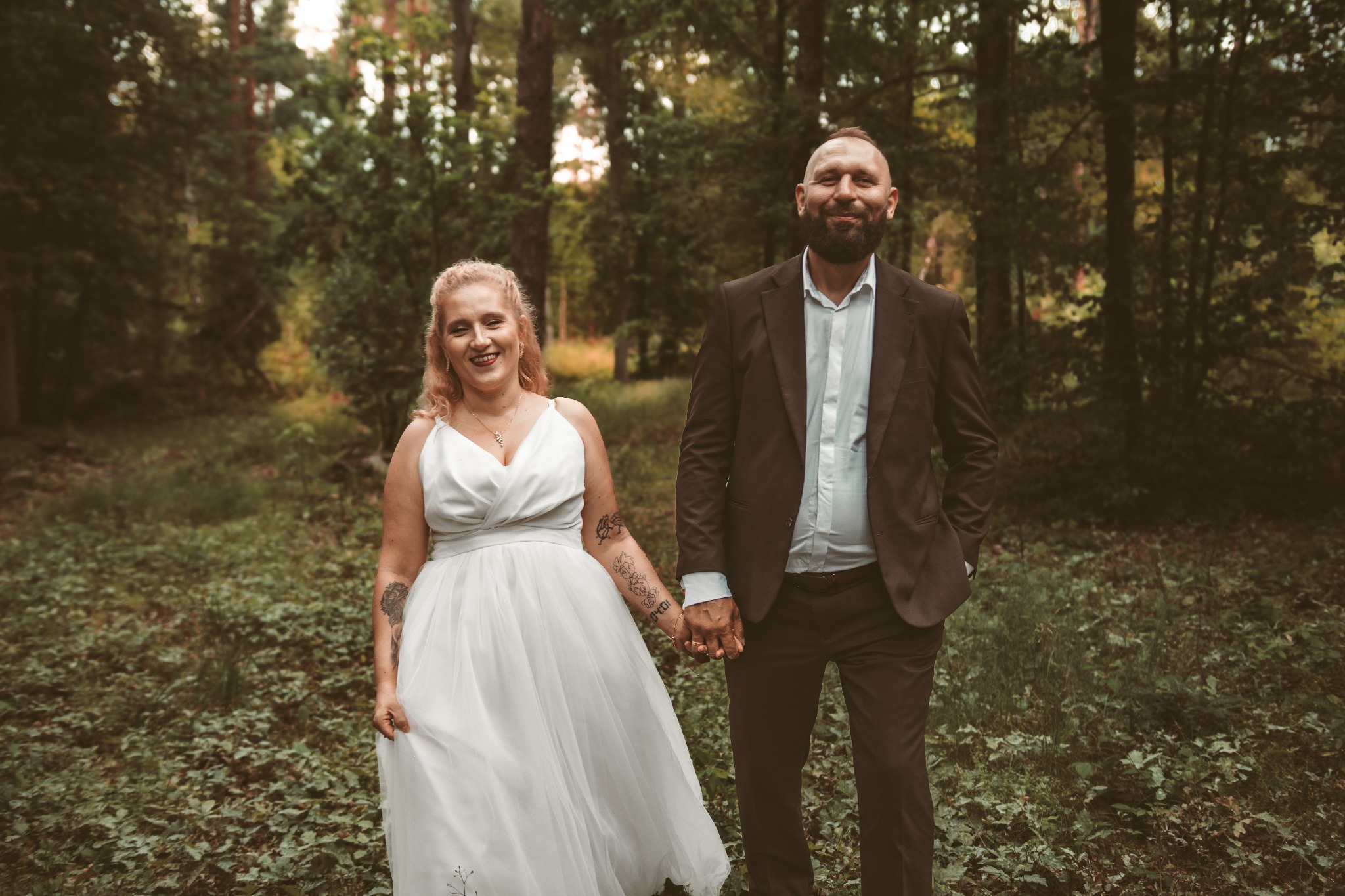 Uśmiechnięta para młoda, trzymająca się za ręce, pozuje na tle leśnej scenerii. Panna młoda w białej sukni z tatuażami na ramieniu, pan młody w brązowym garniturze.