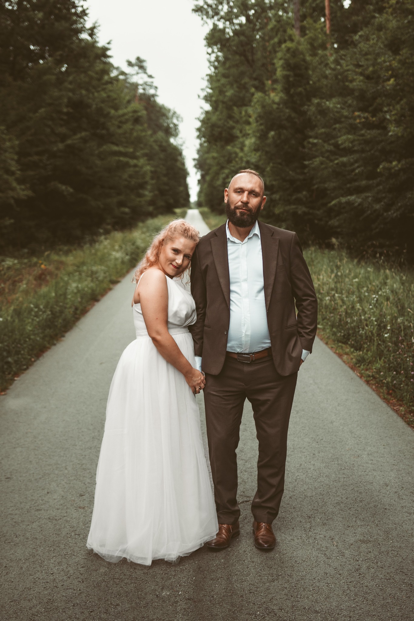 Para młoda na leśnej drodze: panna młoda w białej sukni, pan młody w brązowym garniturze. Romantyczny kadr z perspektywą zbiegającą się w oddali.