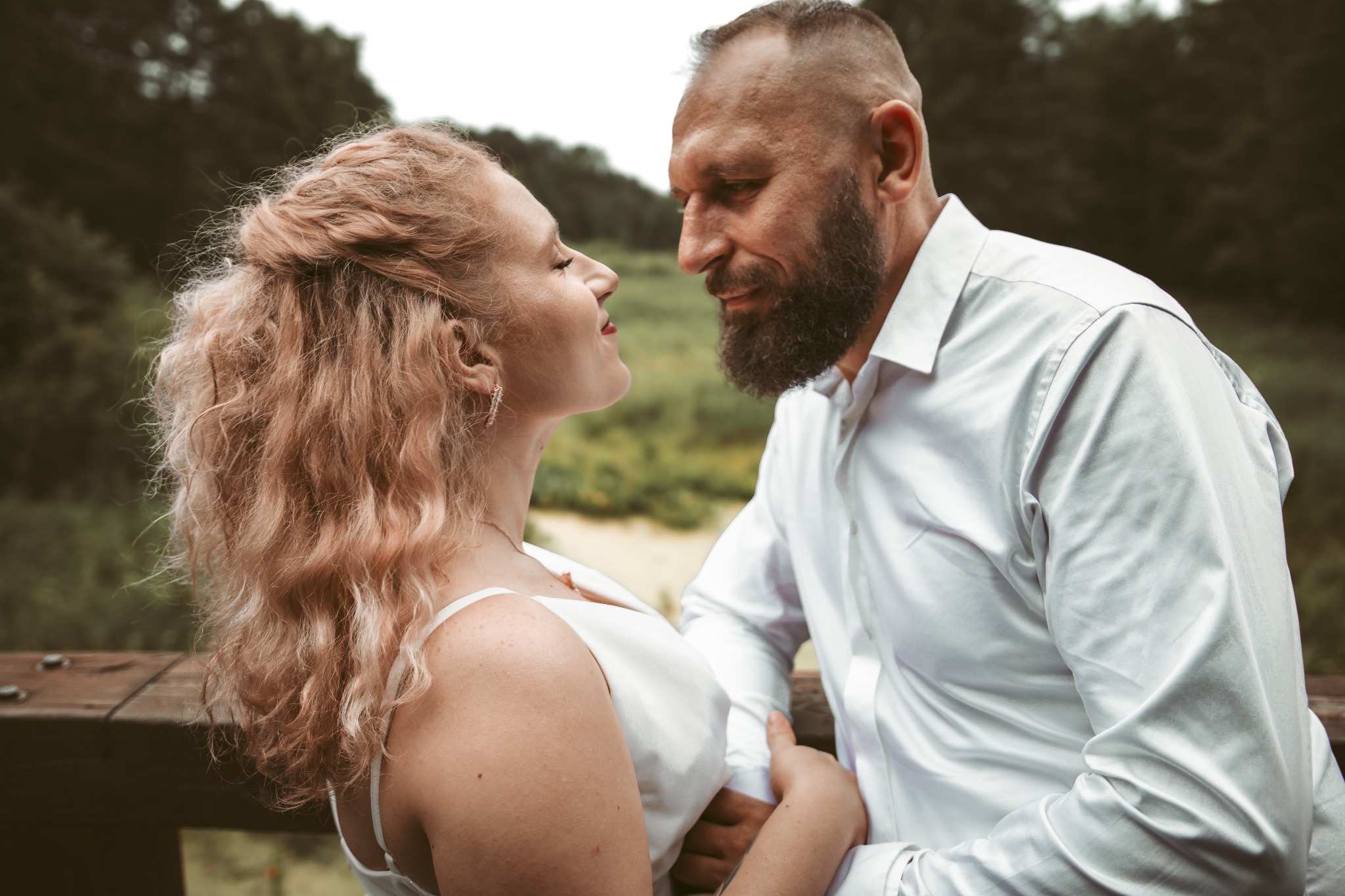 Para zakochanych patrzy na siebie z bliska na tle zieleni. Kobieta ma blond włosy i białą sukienkę, mężczyzna brodę i białą koszulę. Romantyczna sesja w plenerze.