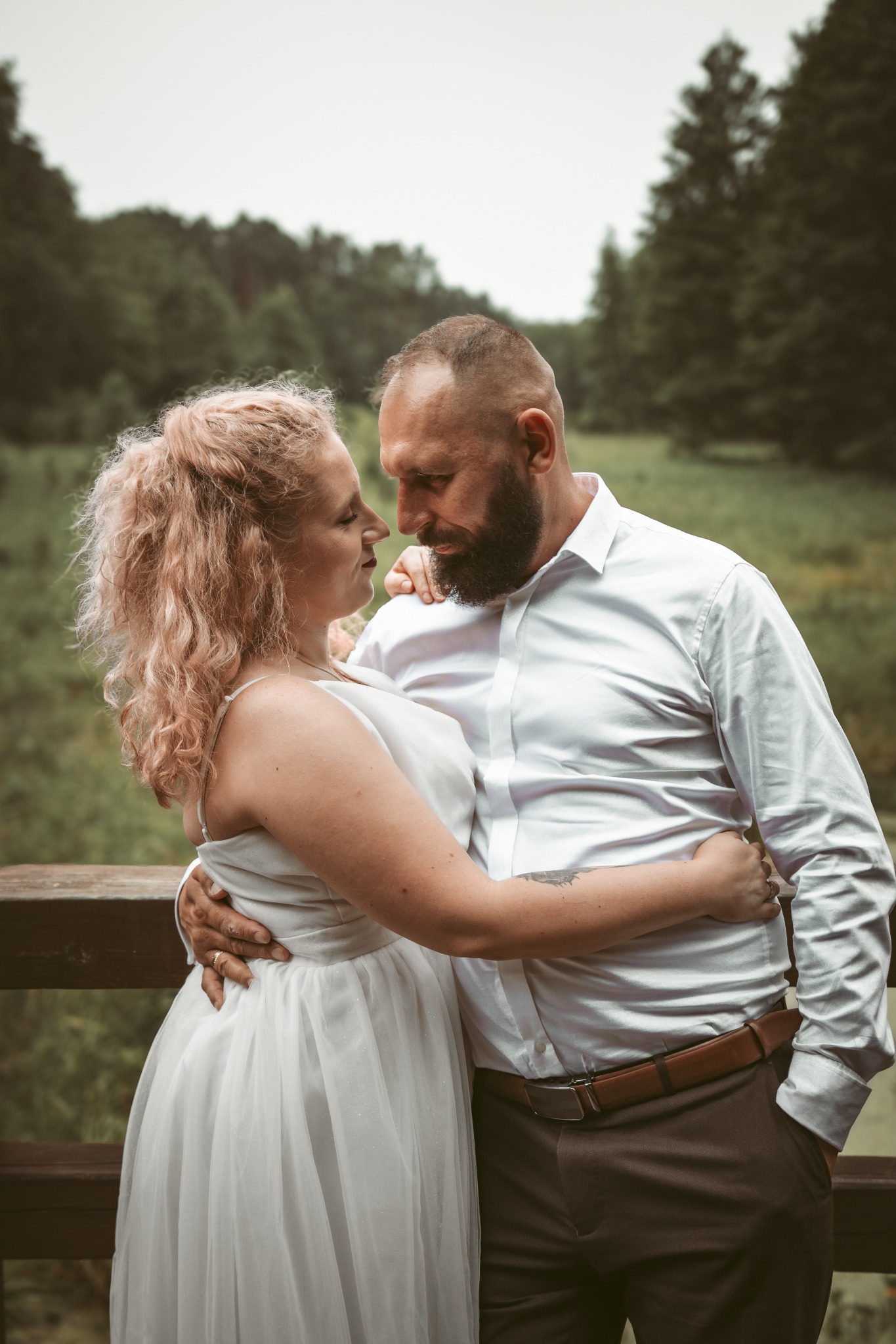 Para w objęciach na tle zieleni. Kobieta w białej sukience, mężczyzna w białej koszuli i brązowych spodniach. Romantyczna sesja w plenerze, delikatne oświetlenie.