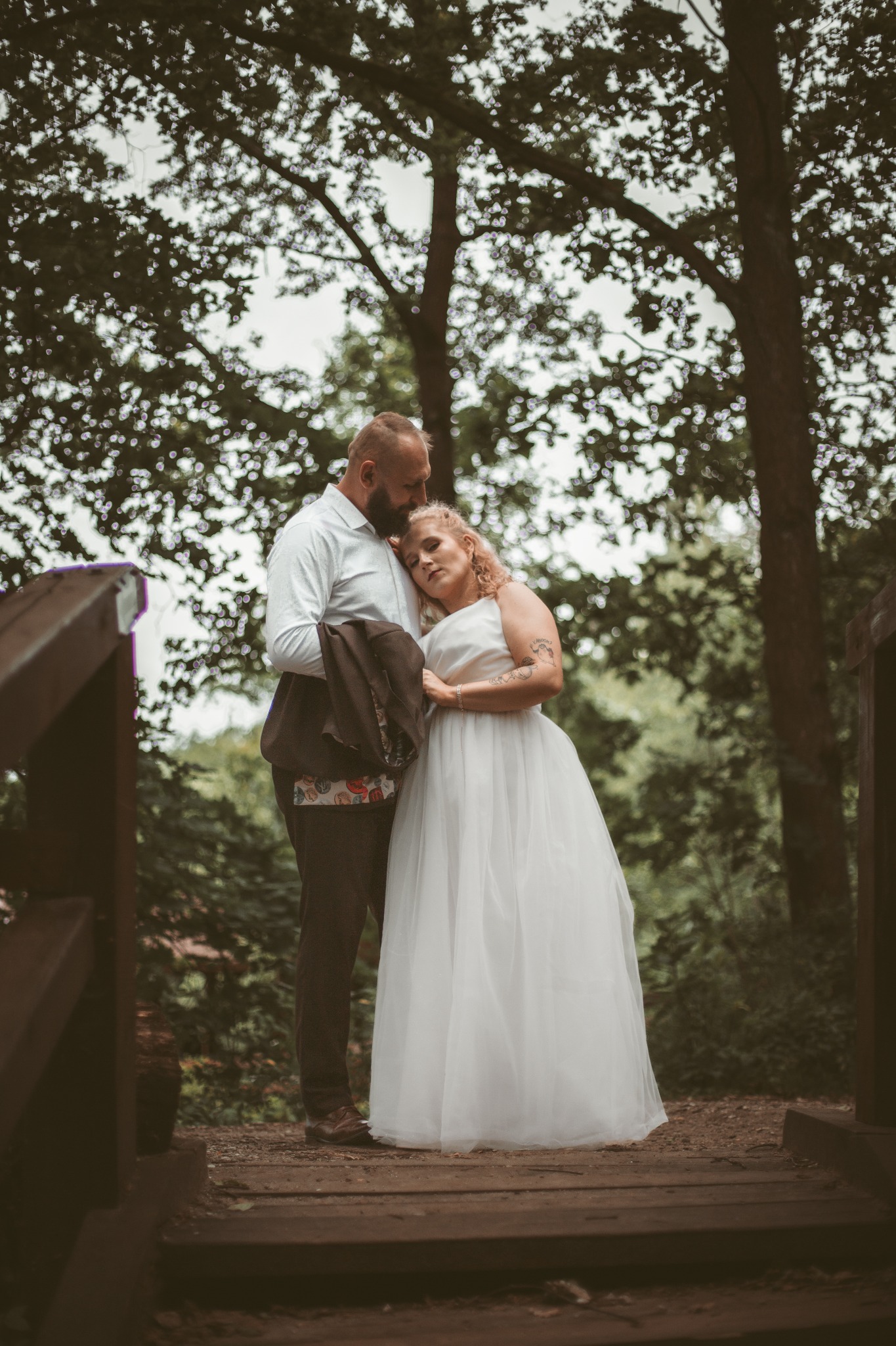 Para młoda w lesie na drewnianym moście. Panna młoda w białej sukni opiera głowę o ramię pana młodego, który trzyma marynarkę. Romantyczna sesja plenerowa.