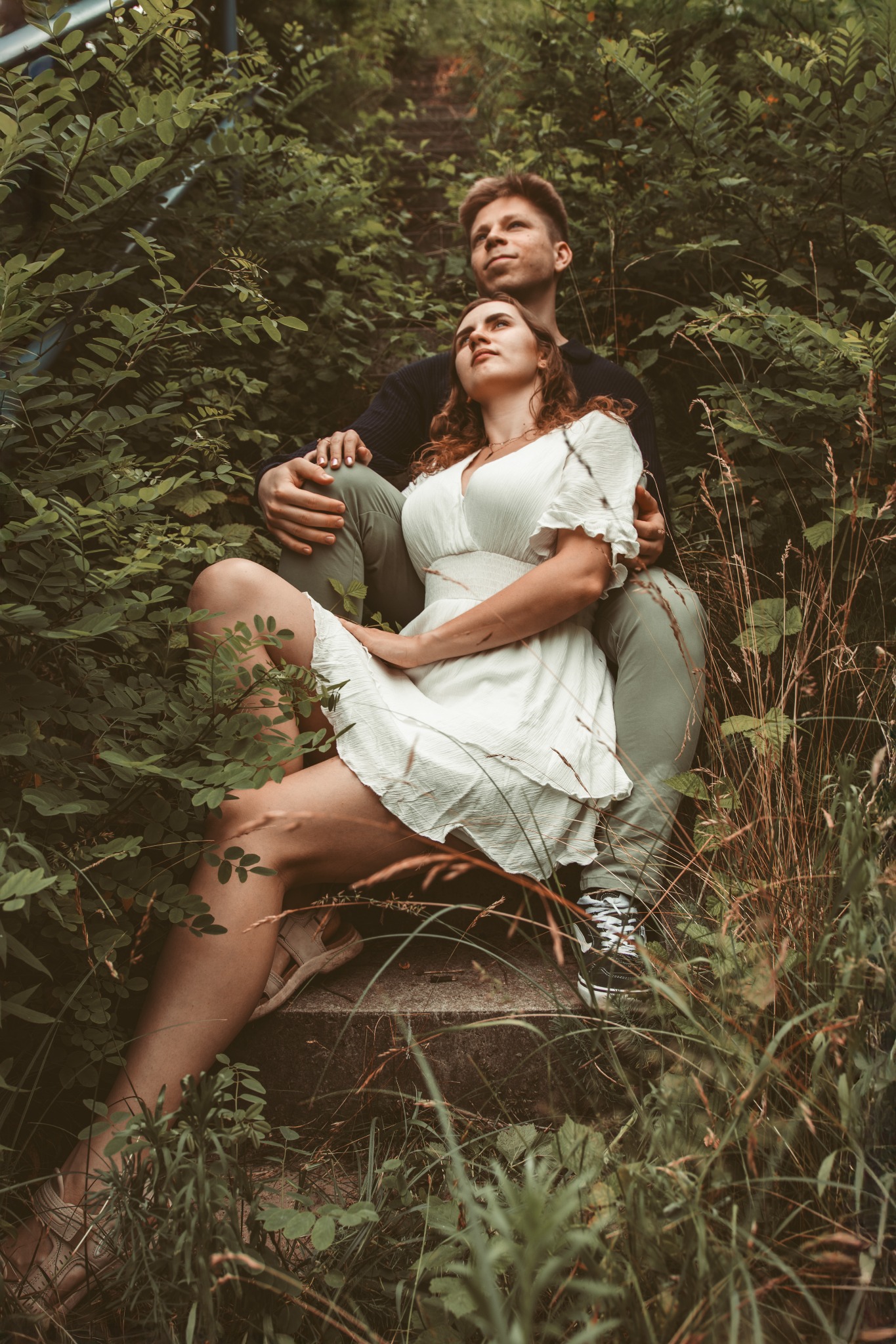 Zakochana para siedzi na betonowych schodach w otoczeniu zieleni. Kobieta w białej sukience, mężczyzna w oliwkowych spodniach. Romantyczna sesja w plenerze.
