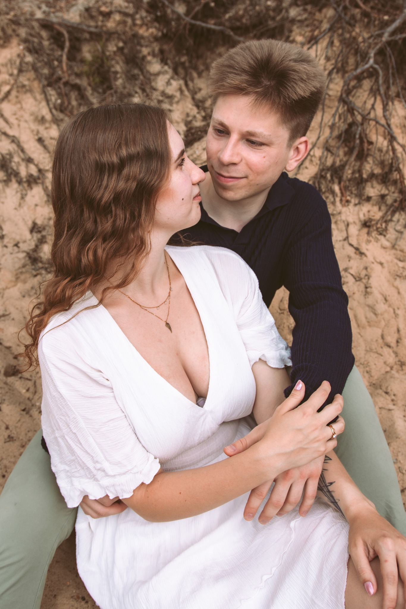 Zakochana para siedzi na piasku, objęta. Kobieta w białej sukience z dekoltem, mężczyzna w ciemnej koszuli. Uśmiechy i bliskość, naturalne światło, delikatny makijaż.