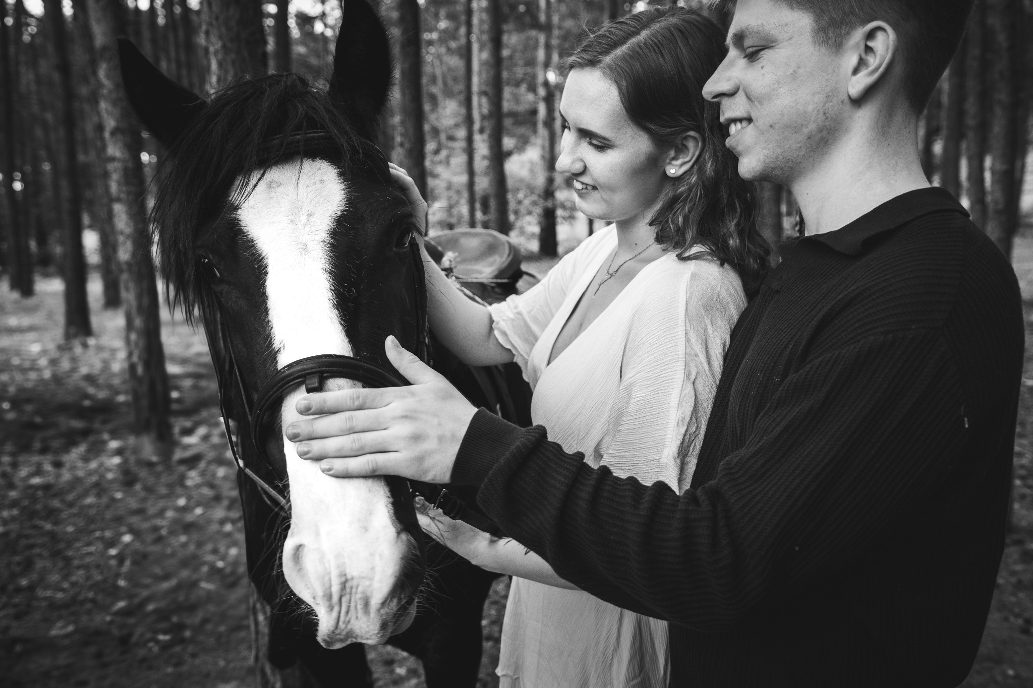 Czarno-białe zdjęcie pary głaszczącej konia w lesie. Kobieta w jasnej sukience, mężczyzna w ciemnym swetrze. Romantyczna sesja w naturalnym otoczeniu.