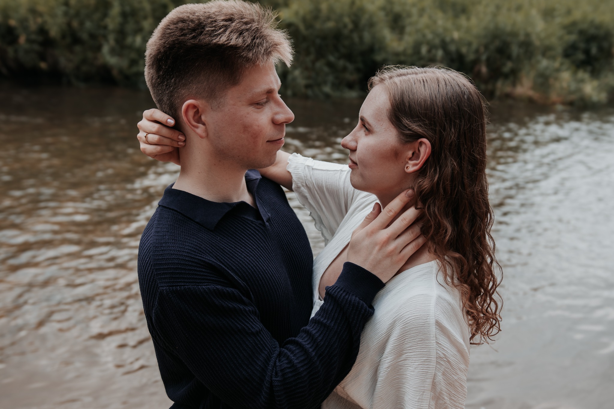 Zakochana para przytulona na tle rzeki, sesja narzeczeńska. Dziewczyna w białej bluzce dotyka szyi chłopaka w granatowym swetrze. Romantyczny plener.