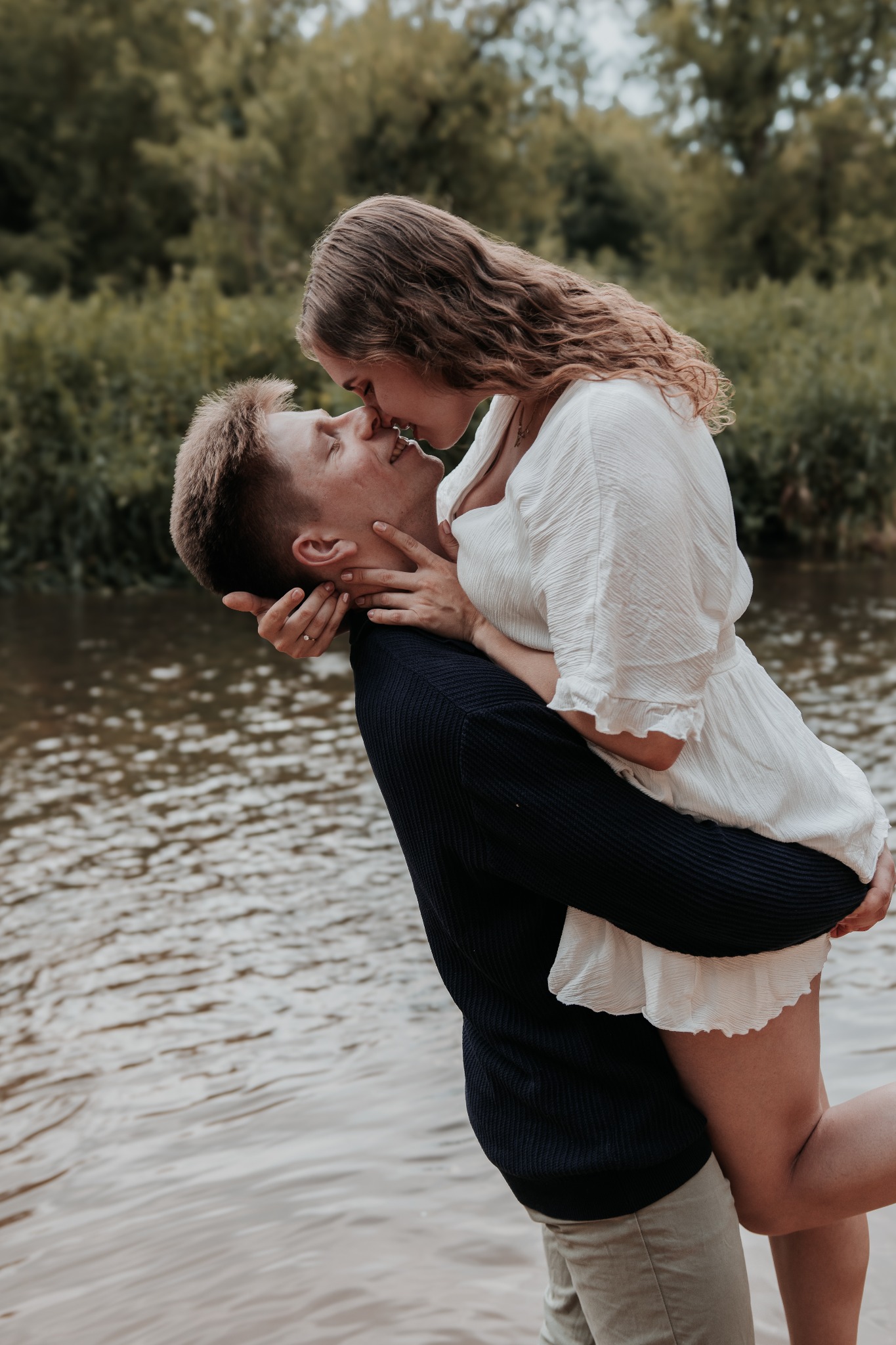 Para zakochanych w romantycznym uścisku na tle rzeki i zieleni. Kobieta w białej sukience, mężczyzna w granatowym swetrze. Zaręczyny symbolizuje pierścionek na palcu.